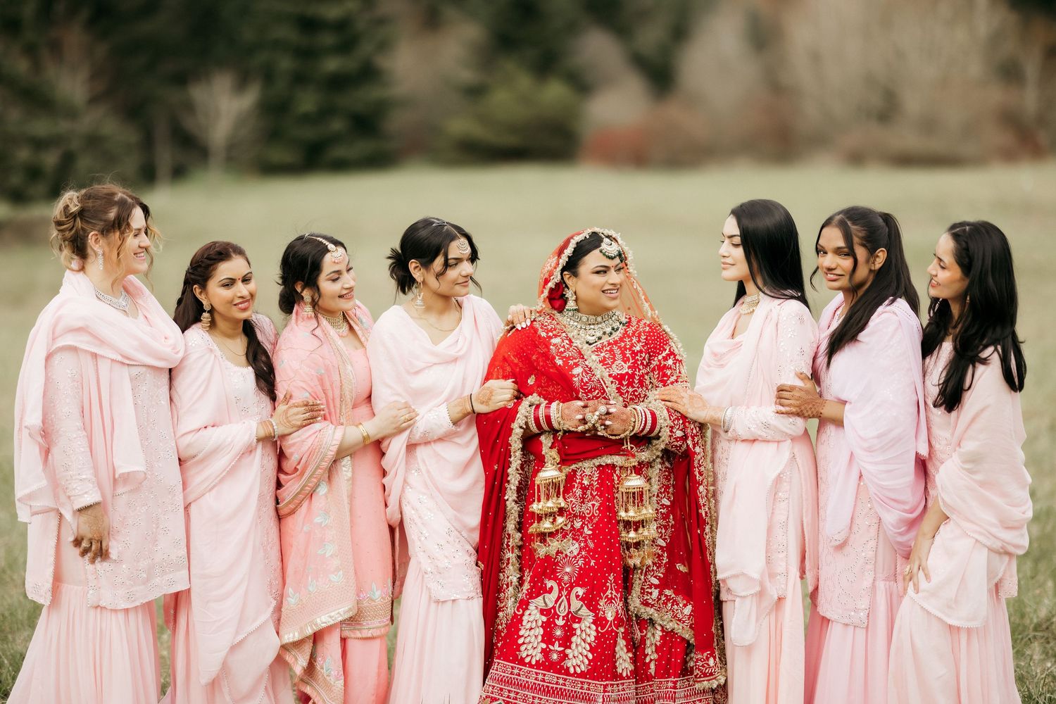 Bridal party in pink outfits gathers around bride in red lehenga for outdoor photos.