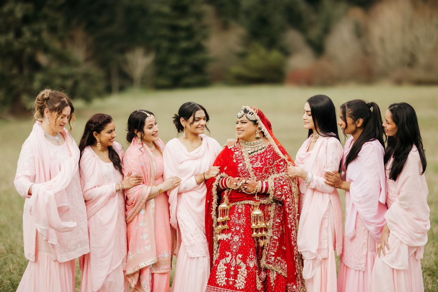 Bridesmaids in pink attire surround bride wearing traditional red Indian wedding lehenga.
