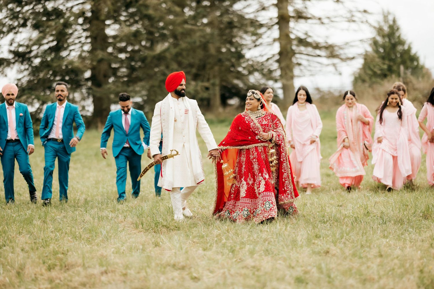 Wedding party in bright colors walks through a grassy field with evergreen trees in background.