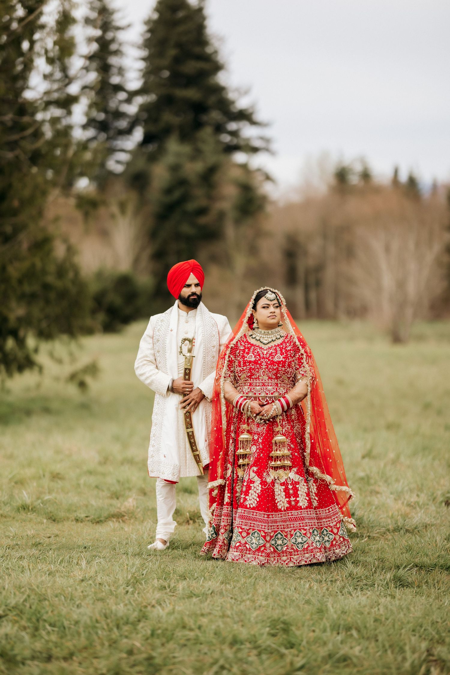Traditional Sikh wedding couple in red and ivory attire standing together in outdoor setting.