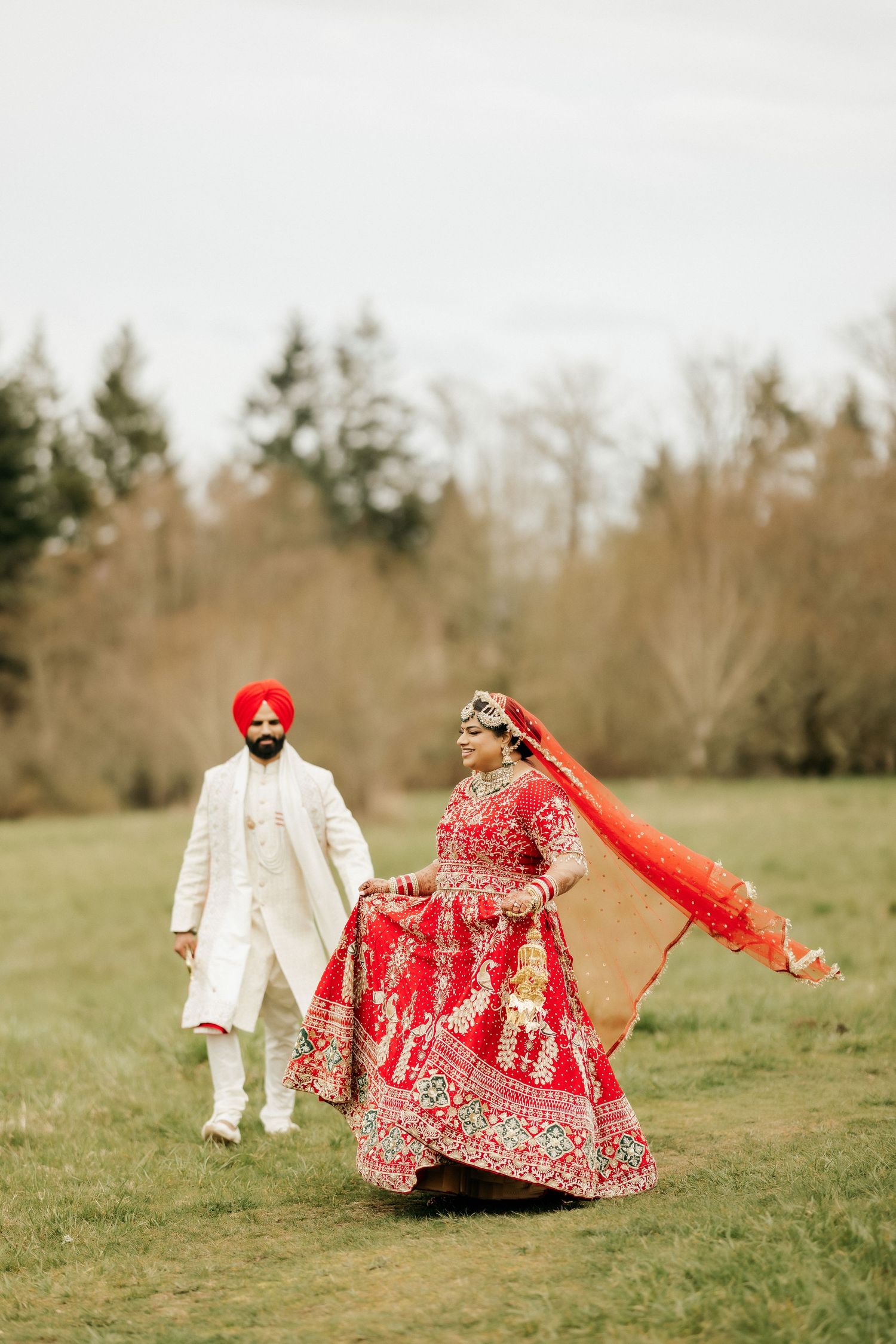 Bride in red lehenga twirling her dupatta while groom watches in outdoor setting.