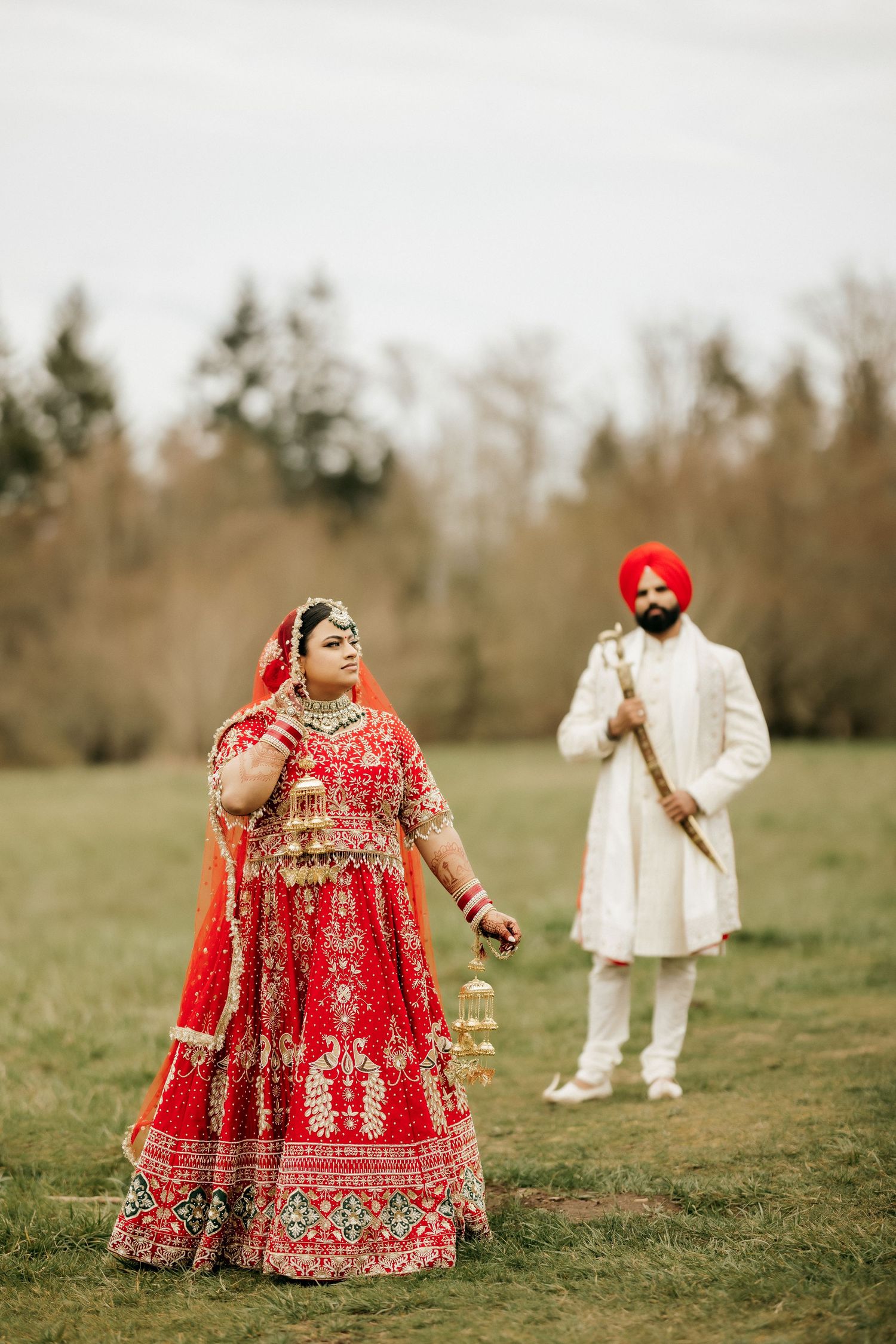 Wedding couple in traditional Sikh attire posing separately in green field with forest background.