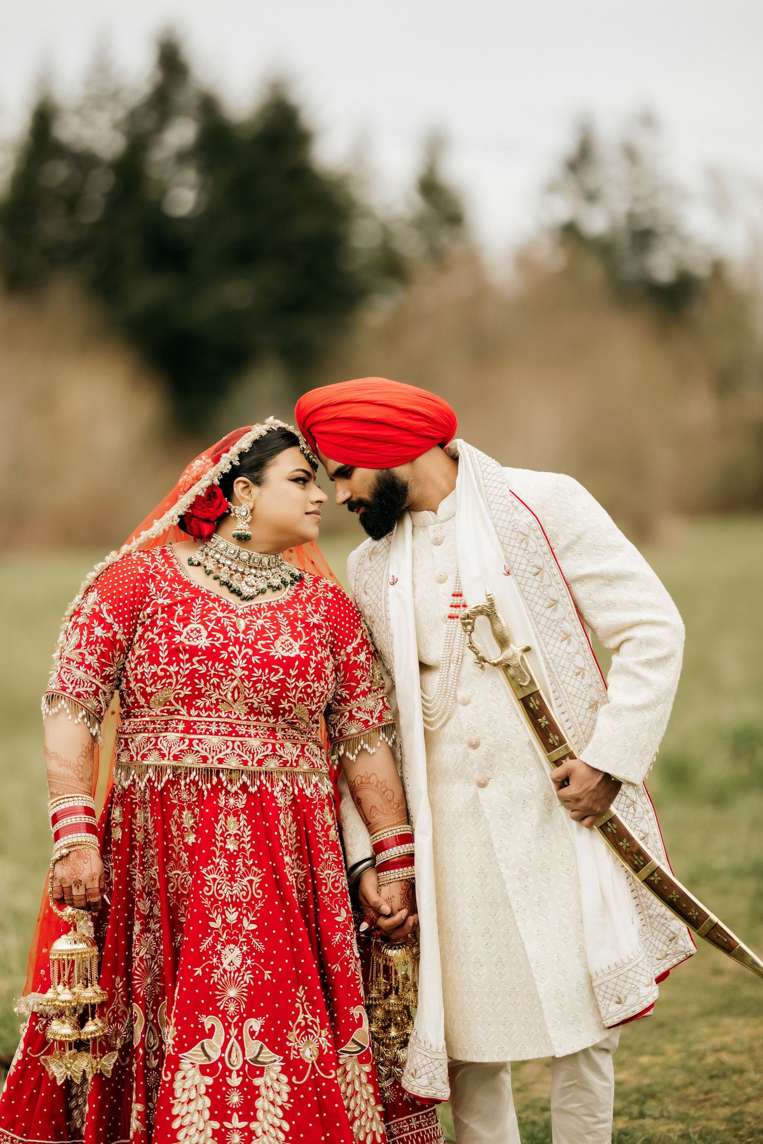Traditional Indian wedding couple in ornate red and ivory outfits sharing intimate moment outdoors.