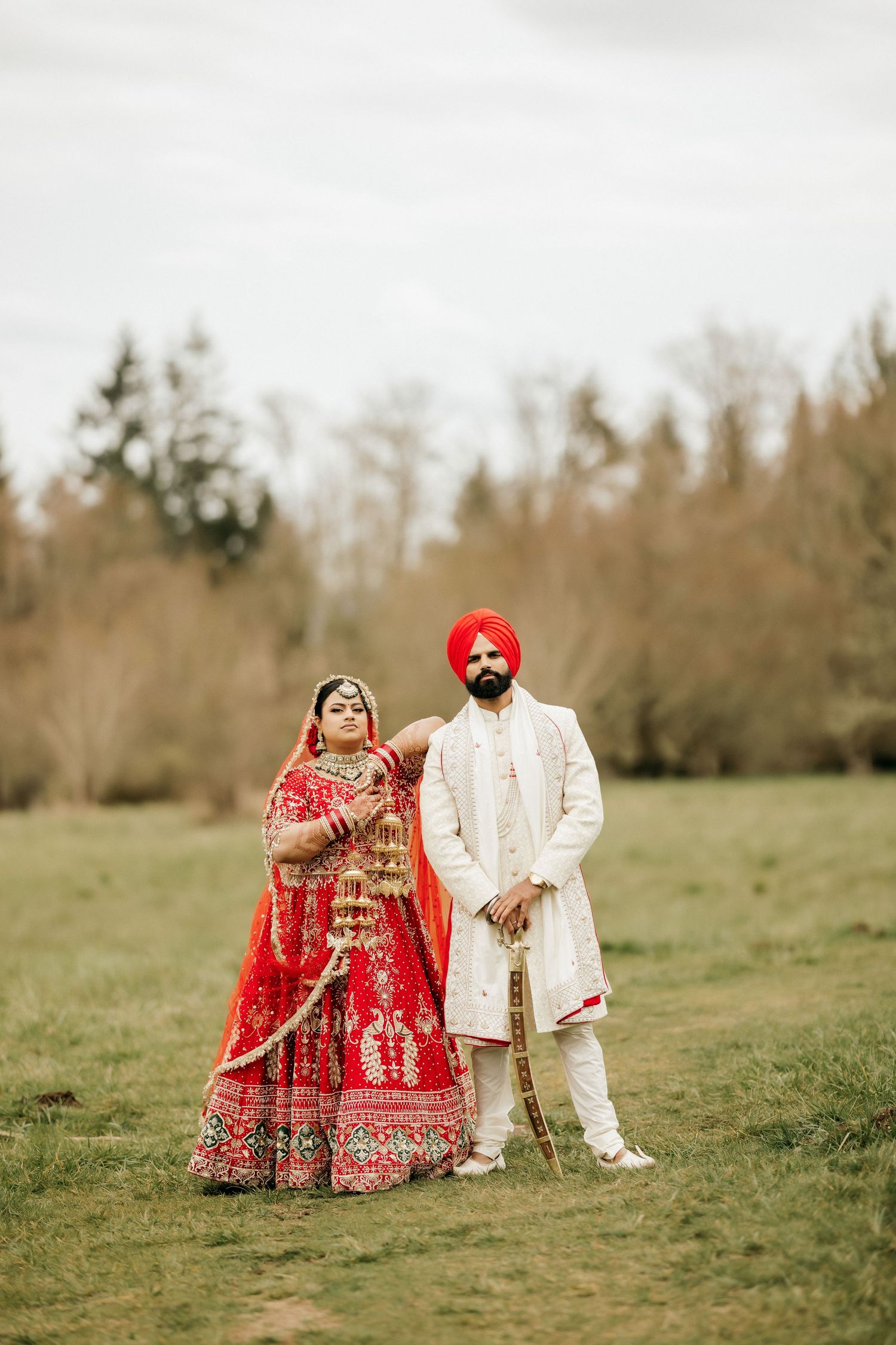 Sikh bride and groom in traditional wedding attire standing in field with forest backdrop.