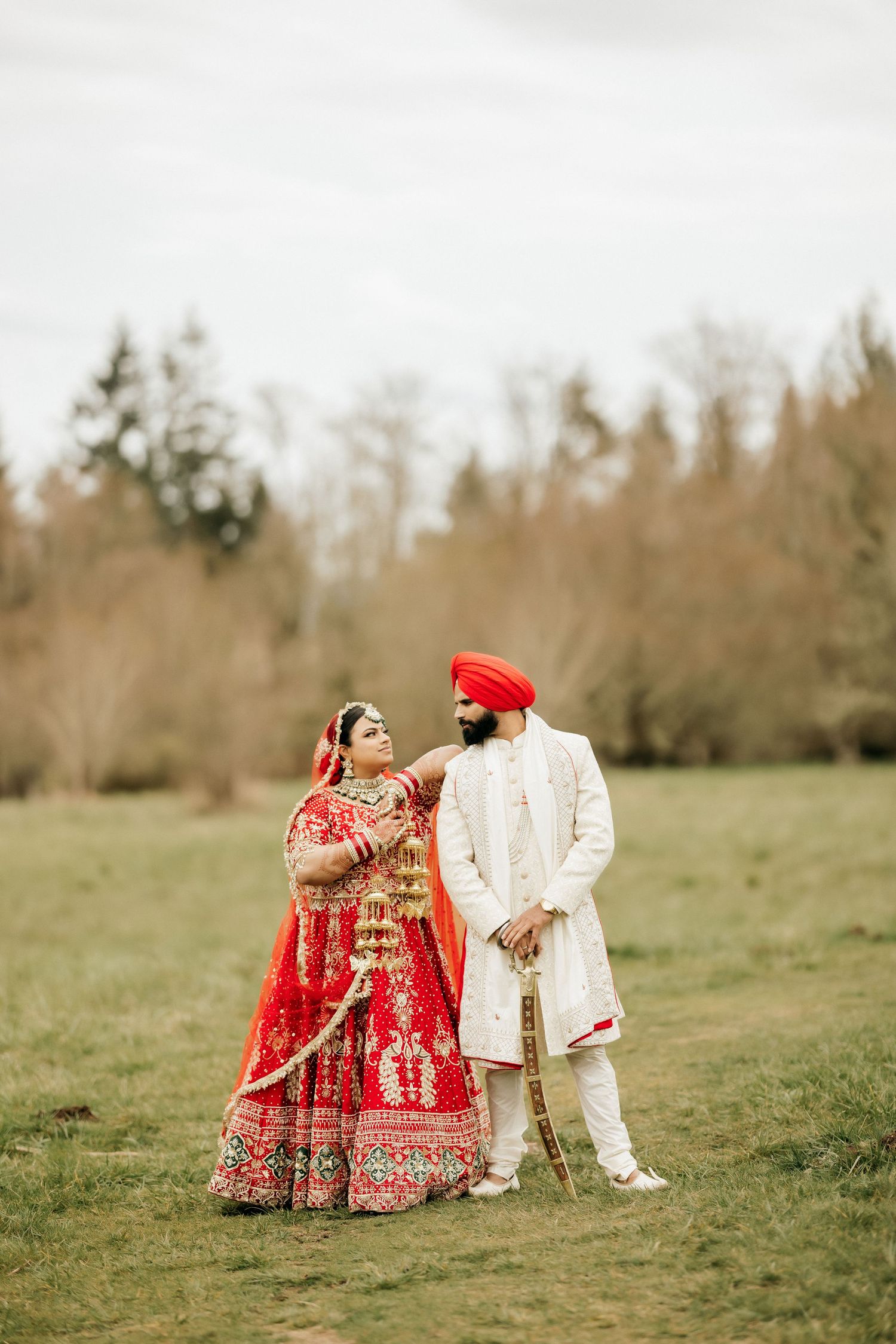 Traditional Indian wedding couple in red and ivory outfits posing together in outdoor setting.