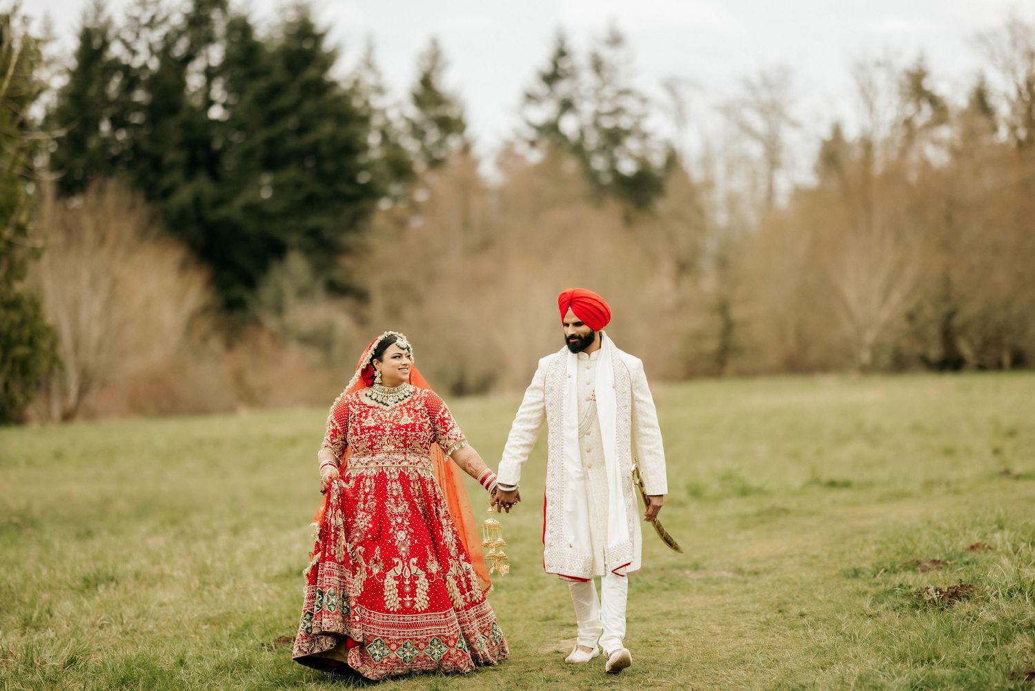 Traditional Sikh couple in wedding attire walking together in a grassy field with evergreen trees in background.