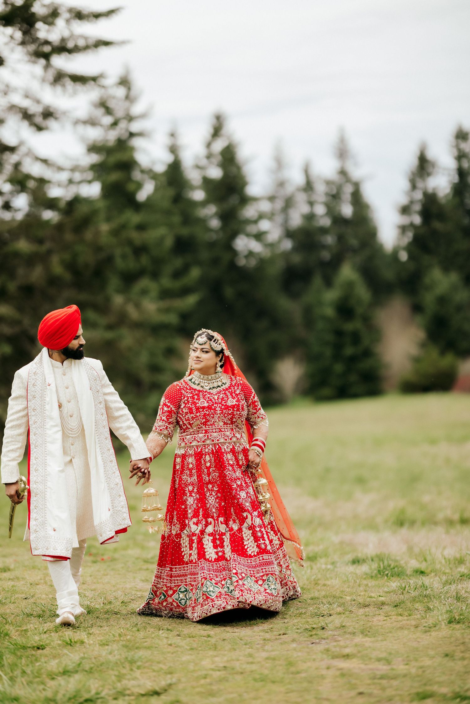 A couple in traditional wedding attire walks hand in hand through an evergreen forest.