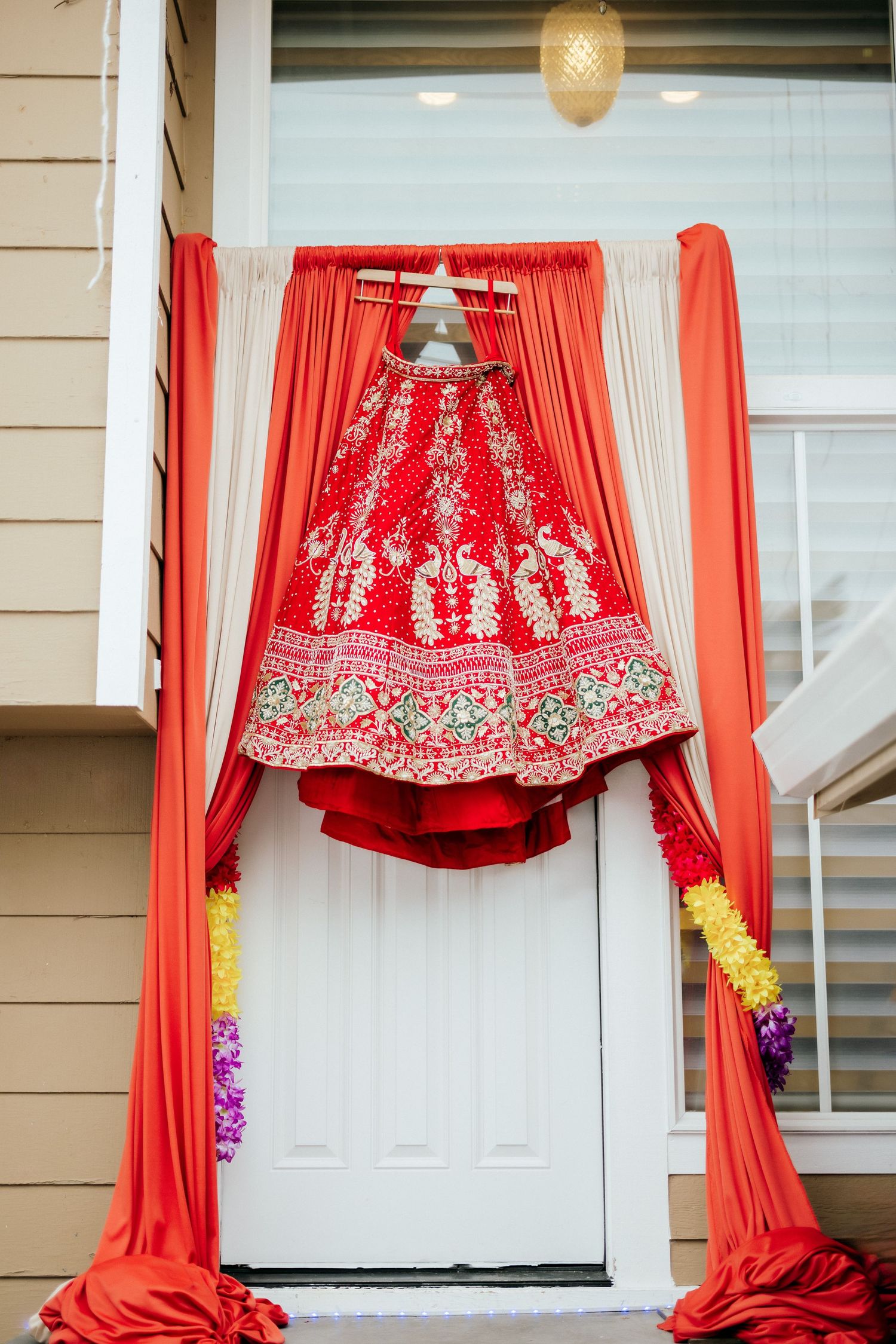 A red and gold embroidered wedding lehenga displayed on a door with draped coral fabric.