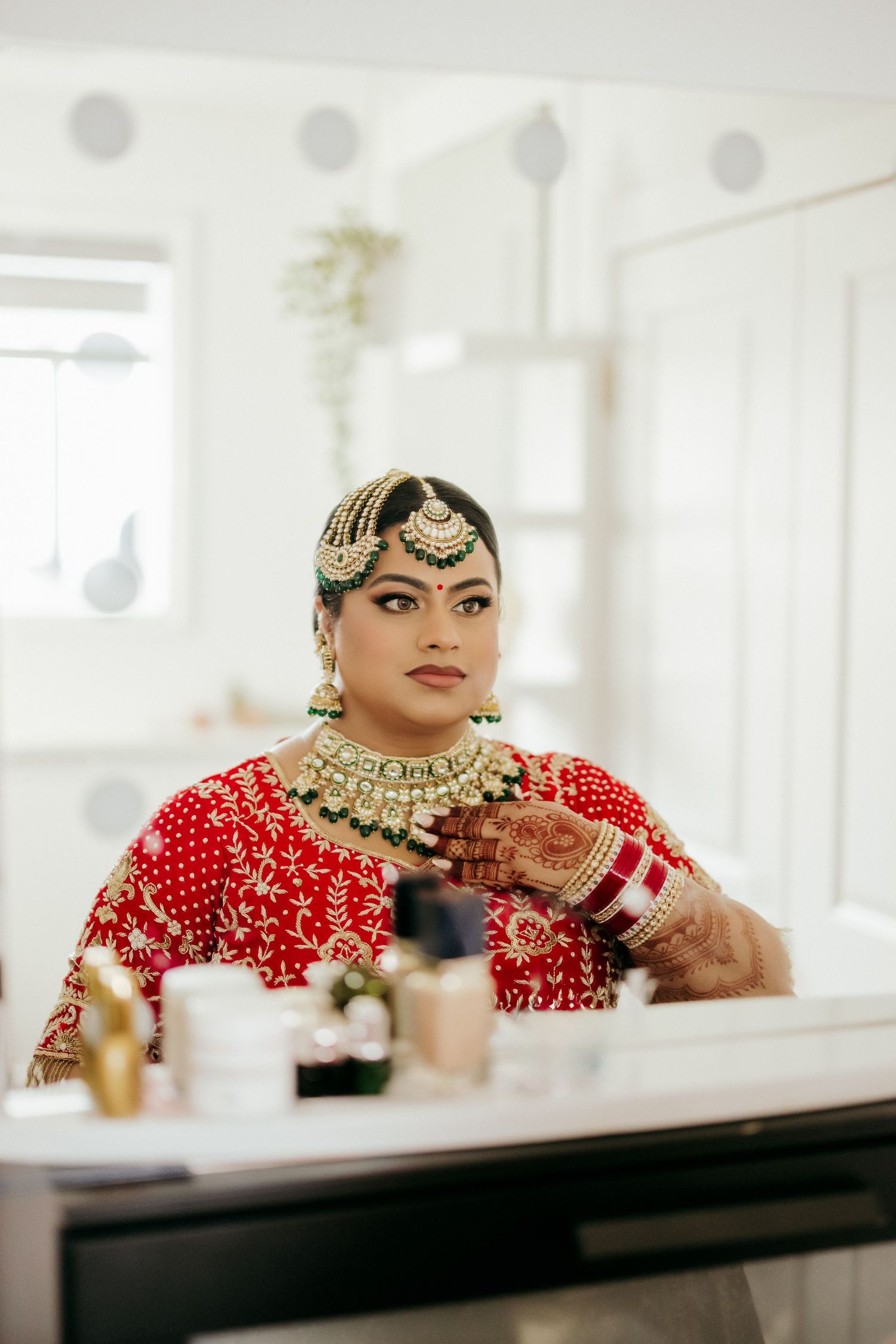Traditional Indian bridal attire with red lehenga, ornate jewelry and henna decorated hands in a bright setting.