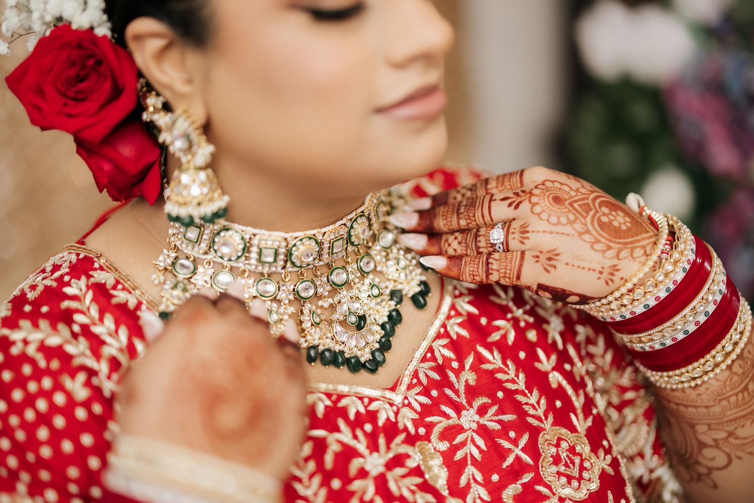 Close-up of traditional Indian bridal jewelry and red lehenga with intricate henna designs and a red rose accent.