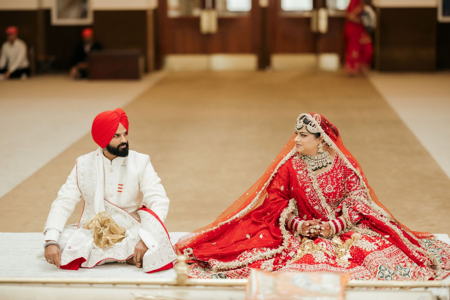 A couple sits on floor during traditional Sikh wedding ceremony in ornate outfits.