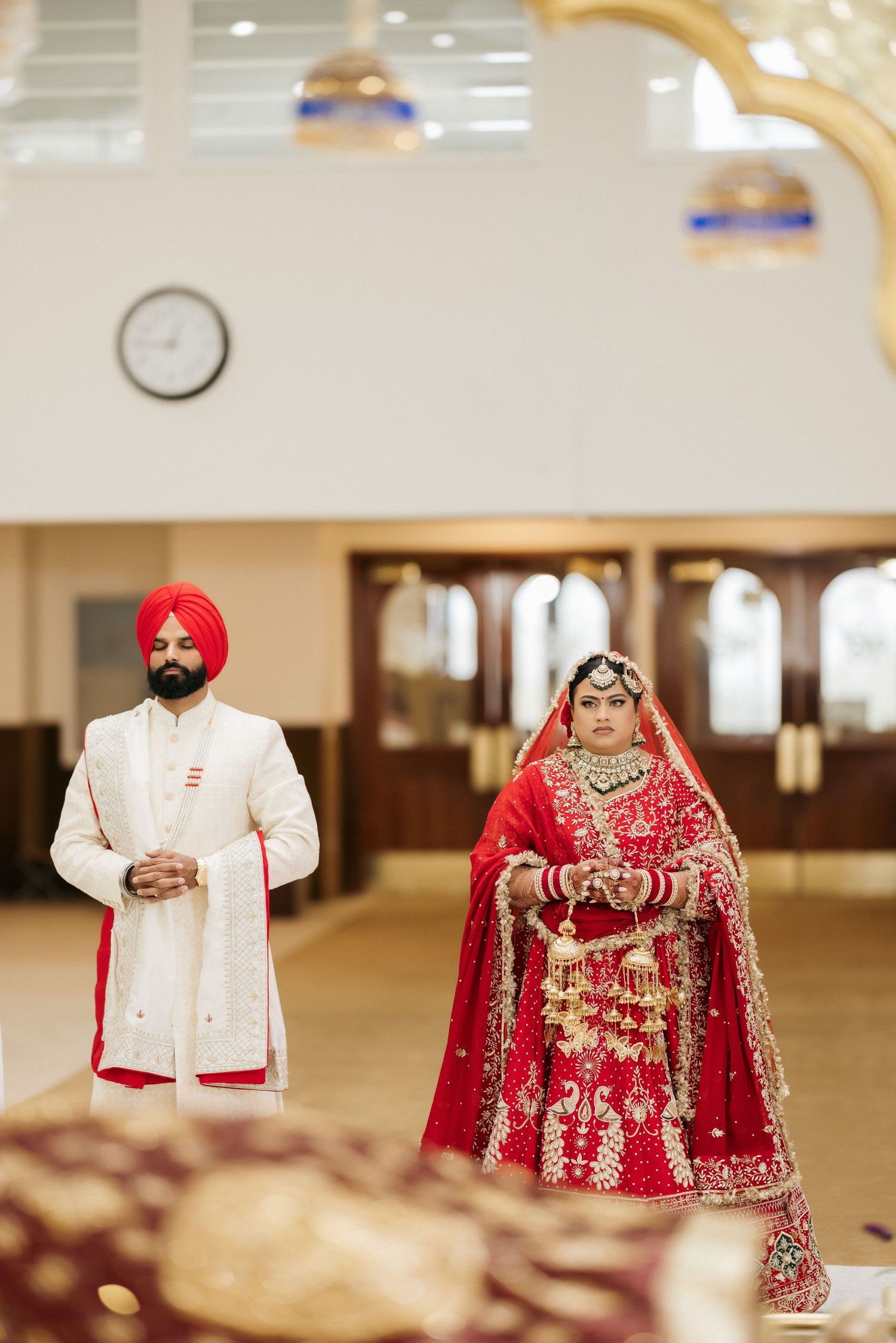 A couple in traditional Sikh wedding attire stands in a temple interior.