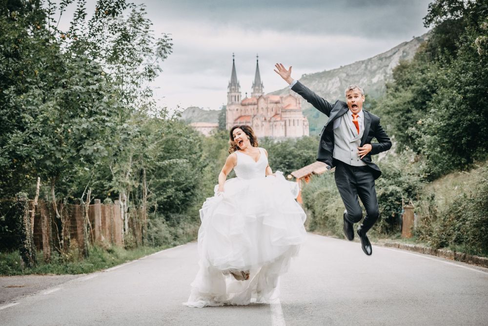 Postboda en Asturias con ambiente desenfadado, pareja corriendo y saltando por una carretera rural con Covadonga al fondo.