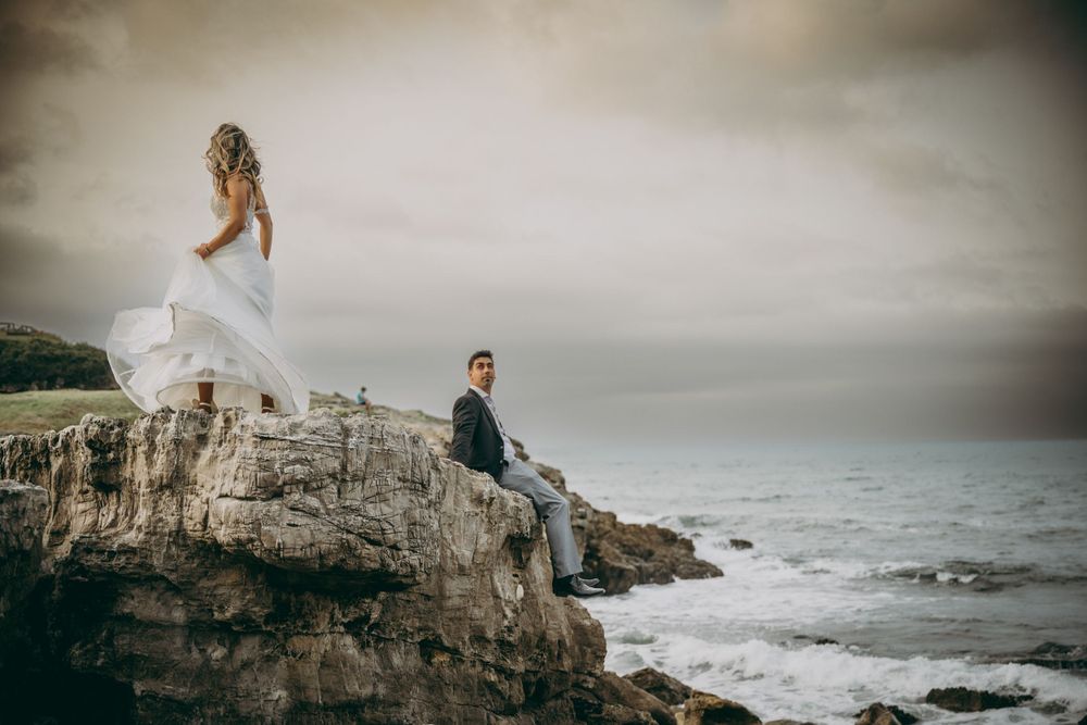 Postboda documental en la costa asturiana, novia sobre una roca frente al mar mientras el novio la observa.