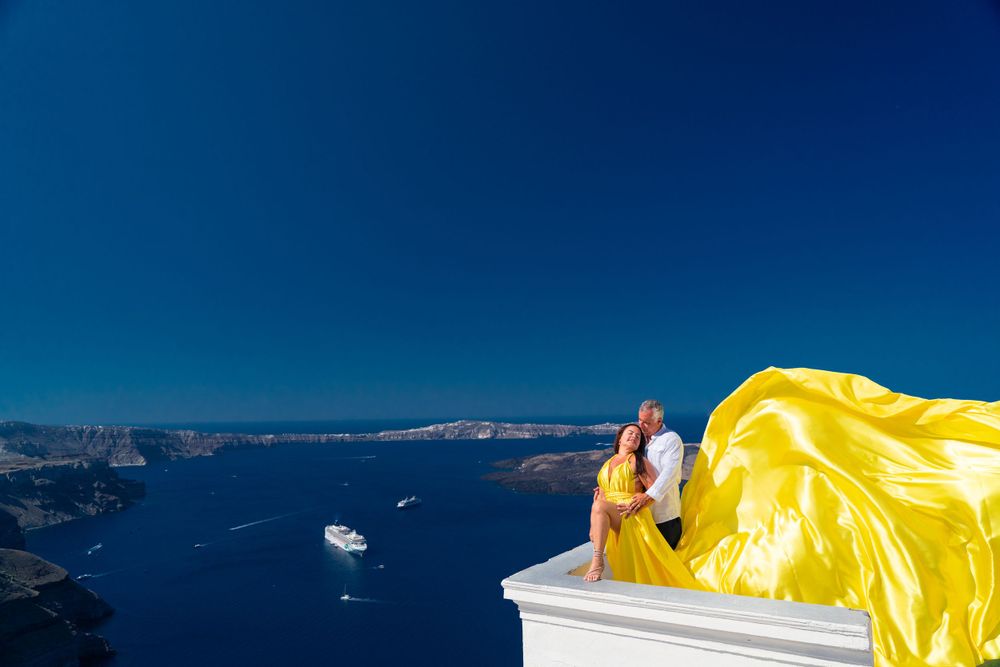 A flowing yellow dress fabric billows dramatically against the blue Santorini coastline and ocean view.