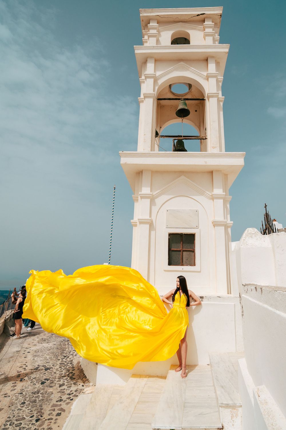 A bright yellow flowing dress fabric billows dramatically against a white bell tower in Santorini, Greece.