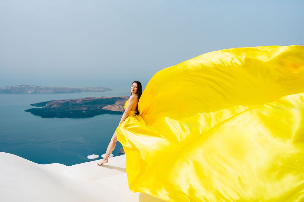 A flowing yellow fabric catches the wind against the backdrop of Santorini's blue waters and white buildings.