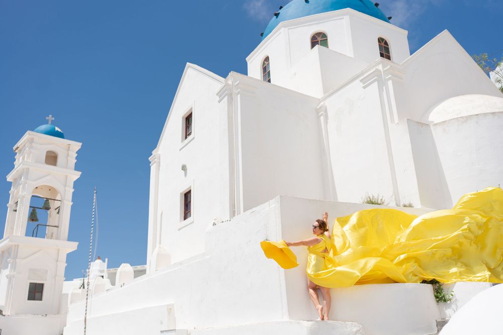 Yellow flowing fabric creates contrast against white church with blue domes in Greek architecture.