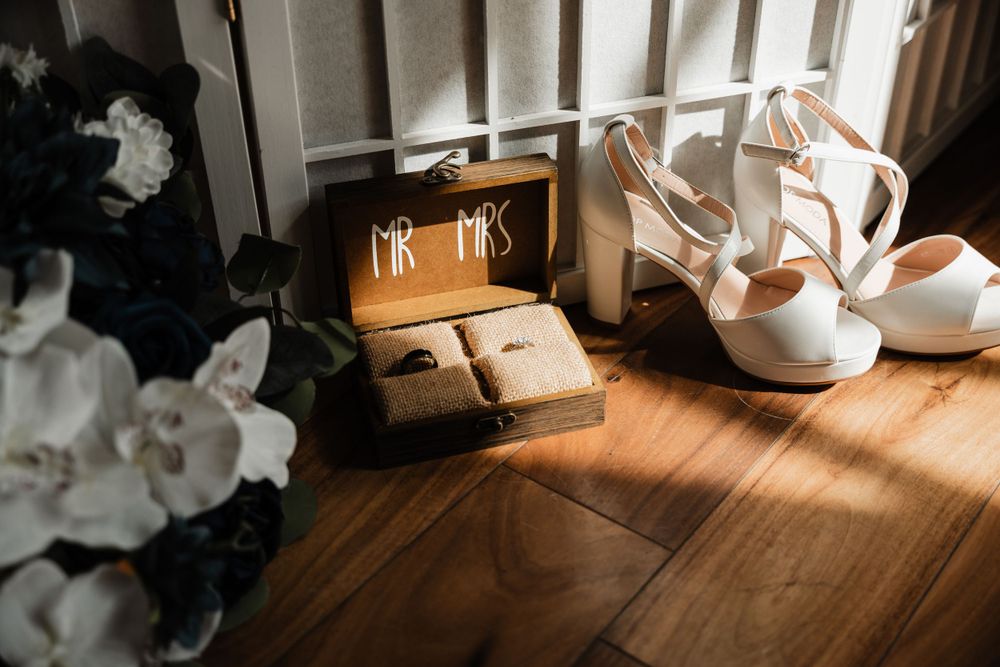 Wedding details including white heels and jewelry boxes on a wooden floor illuminated by natural light.