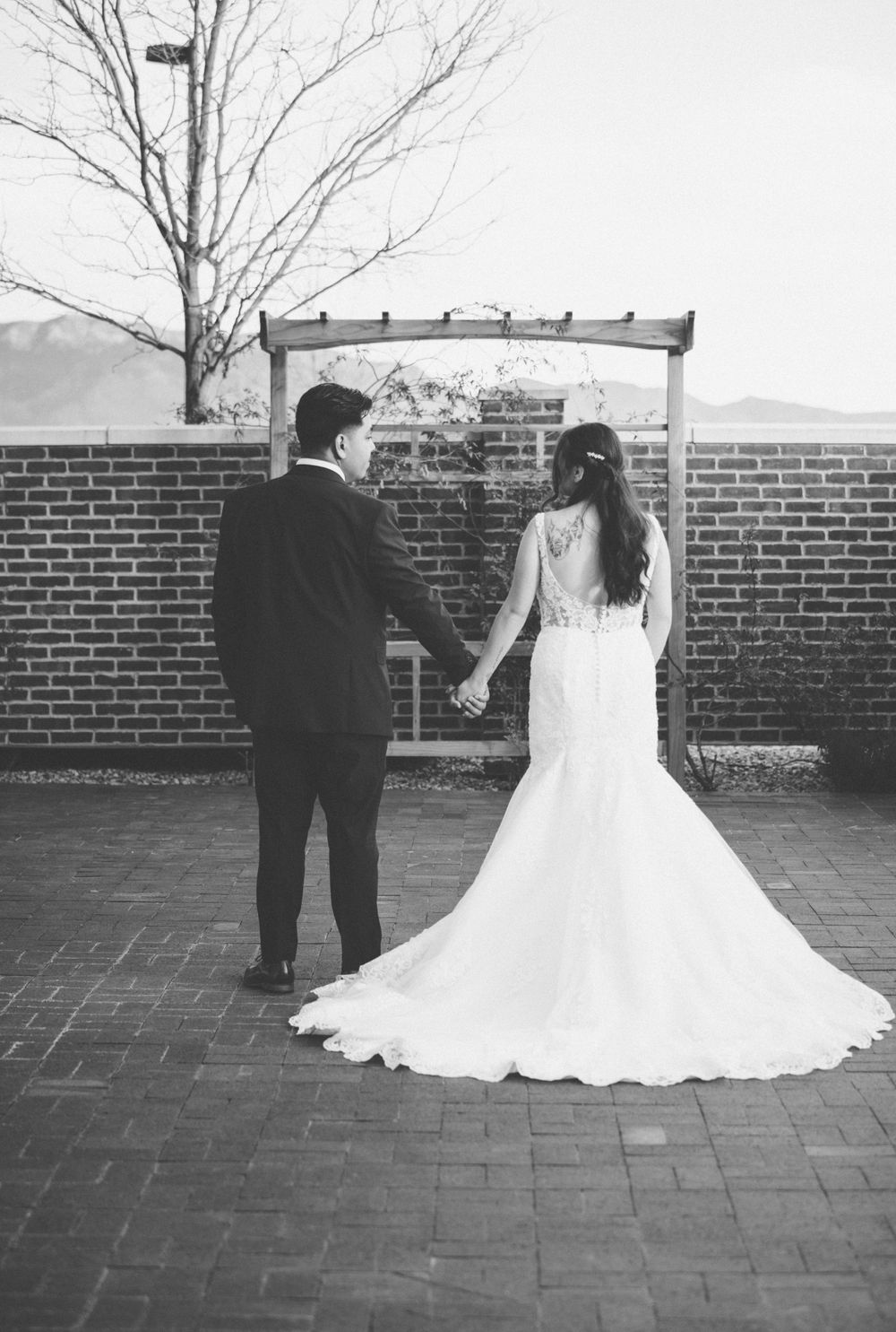 Black and white photo of a couple in wedding attire holding hands near a brick wall.