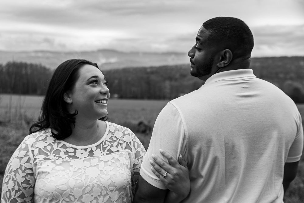 A romantic couple embraces while standing in a field with mountains in the background.