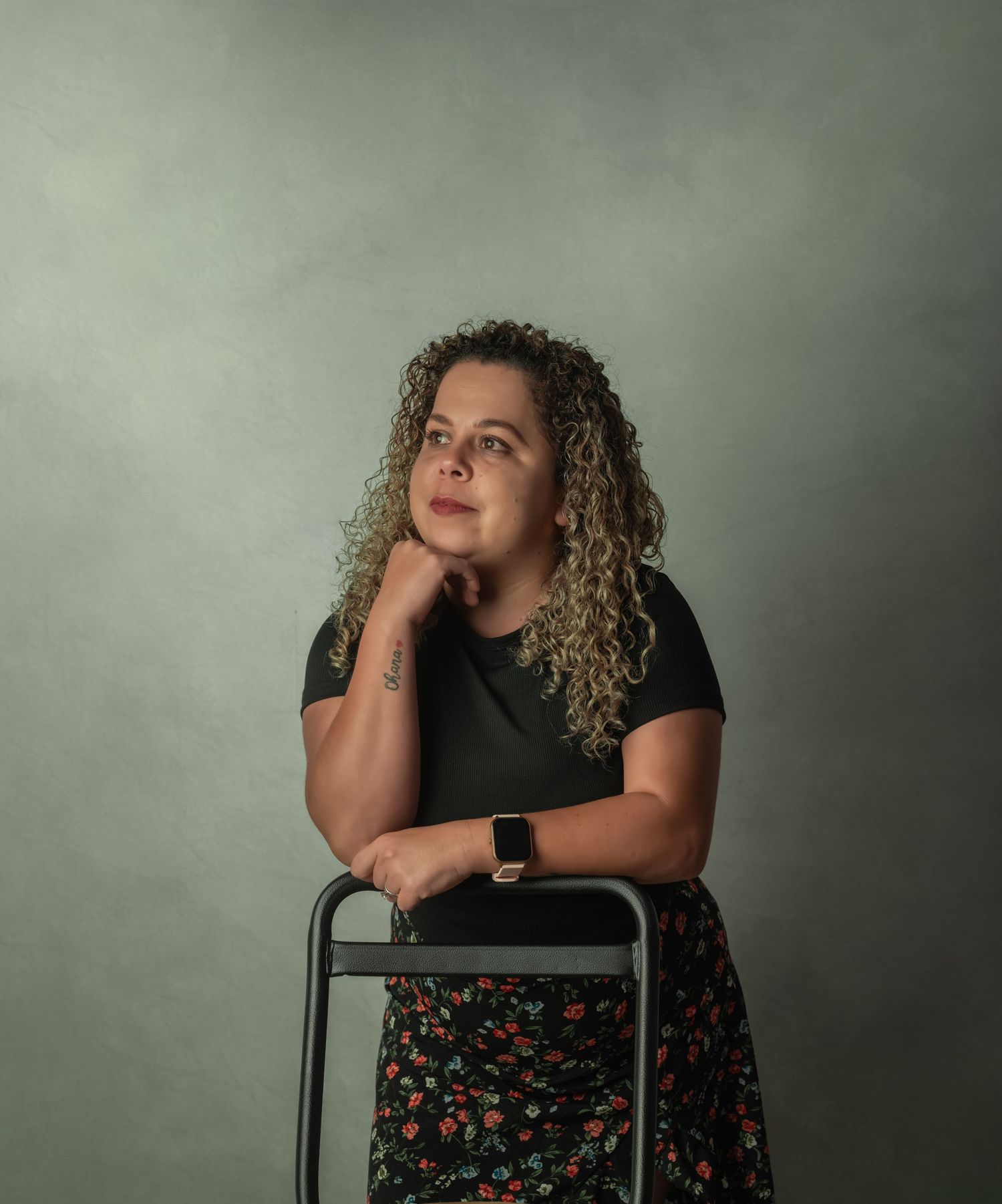 Portrait of a woman with curly blonde hair leaning thoughtfully on a chair against a gray backdrop.