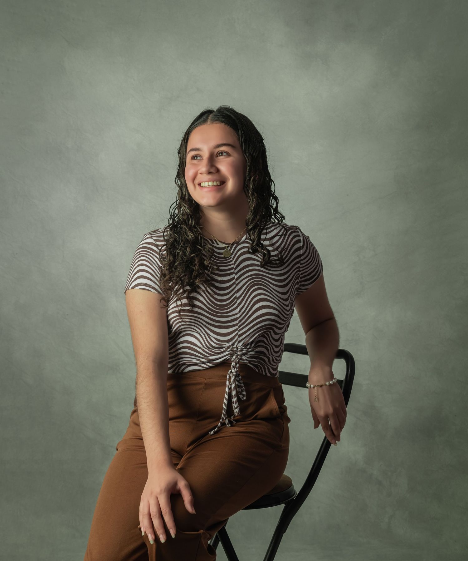 Casual studio portrait of someone in a striped shirt and brown pants seated on a chair.