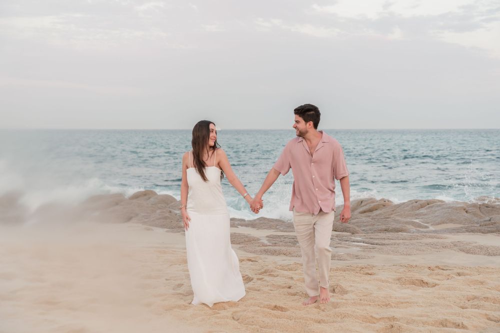 A couple walks hand in hand along a beach shore in romantic sunset lighting.