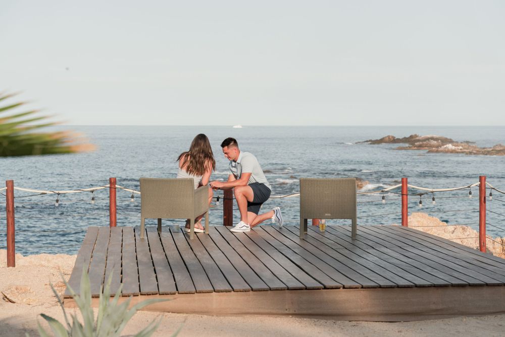 Two people enjoy a romantic moment on a wooden deck overlooking the ocean at sunset.