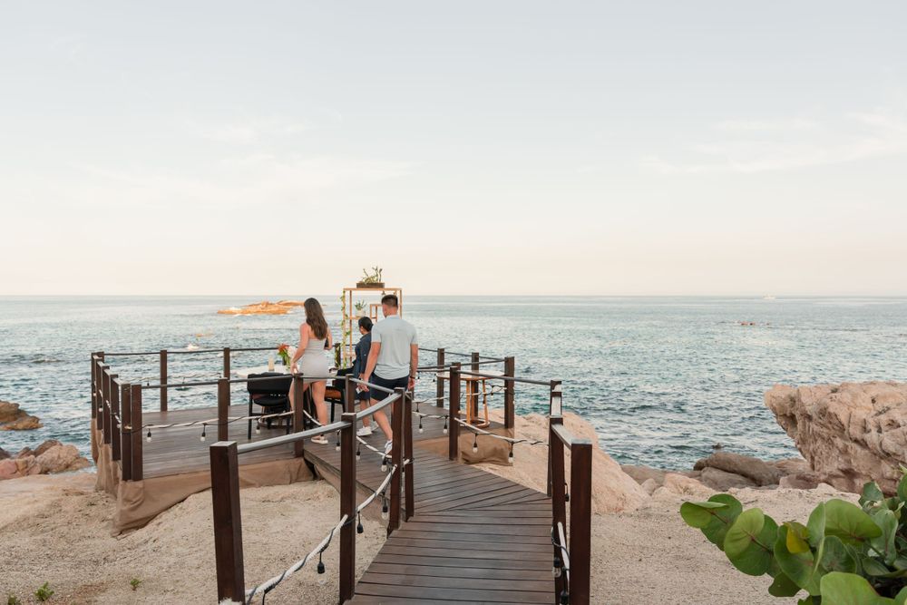 A couple walks along a wooden boardwalk overlooking the rocky coastline at sunset.