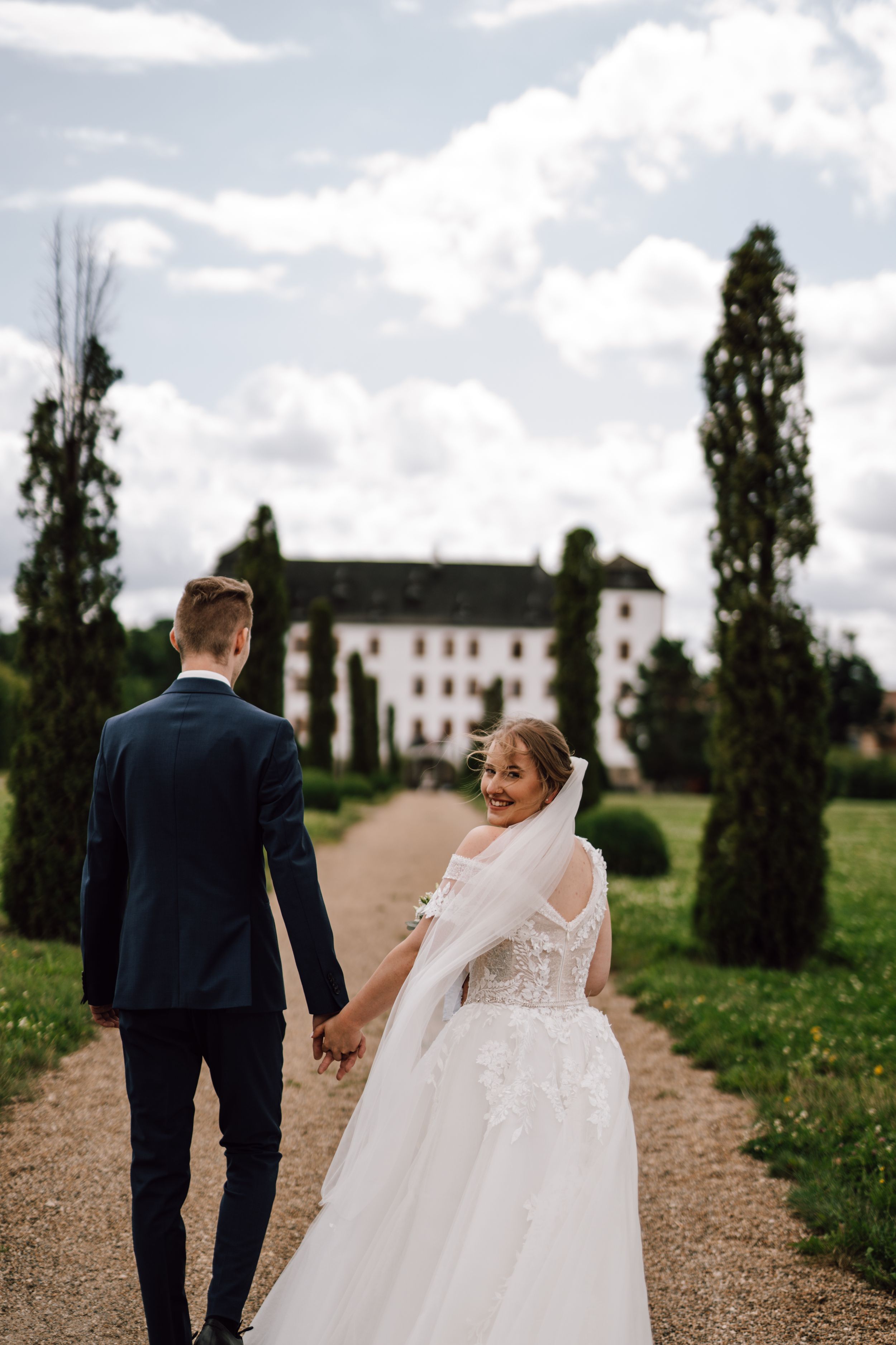 Newlyweds walking down a path towards a stately white manor house with cypress trees.