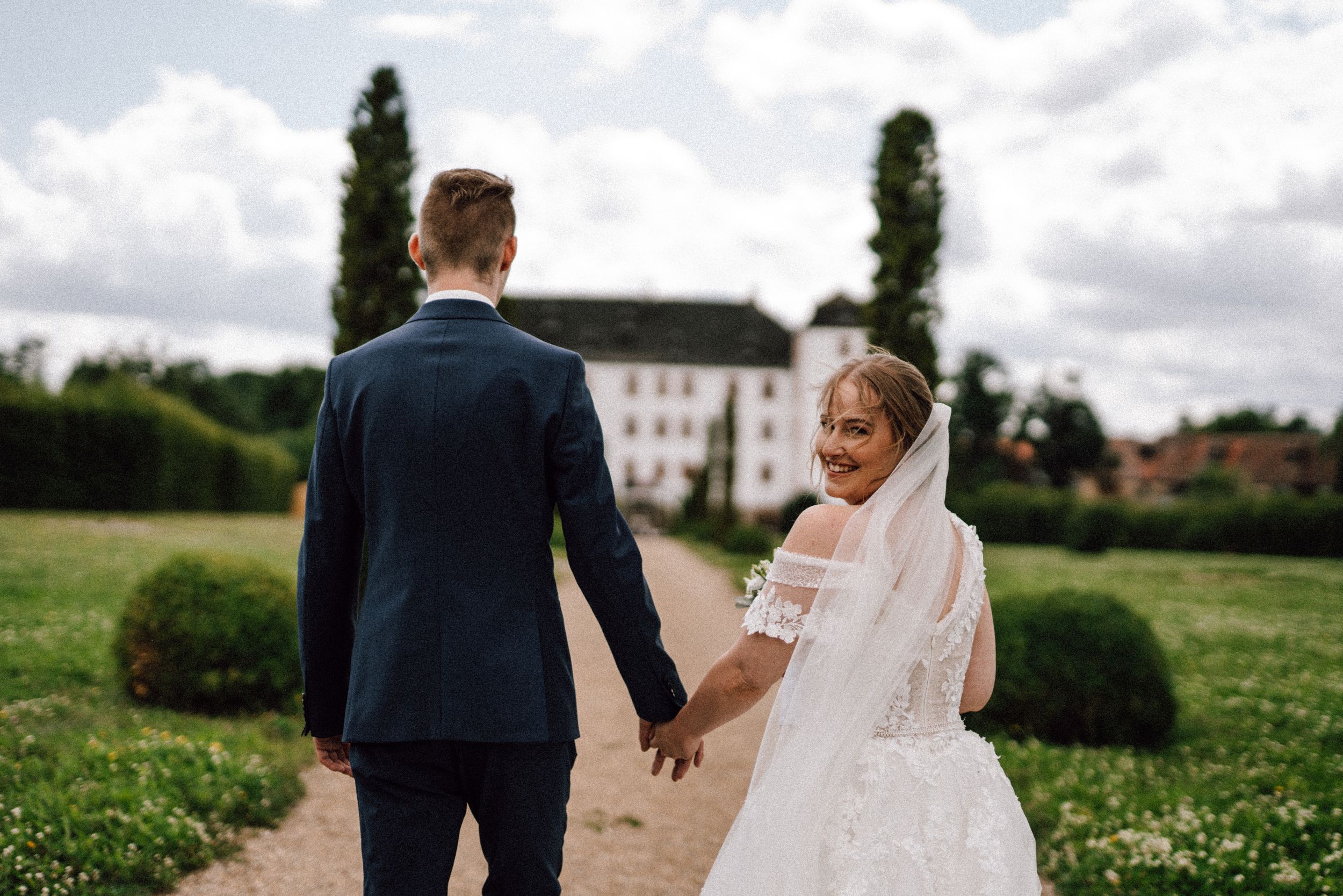 A couple shares a joyful moment on a path leading to an elegant white mansion.