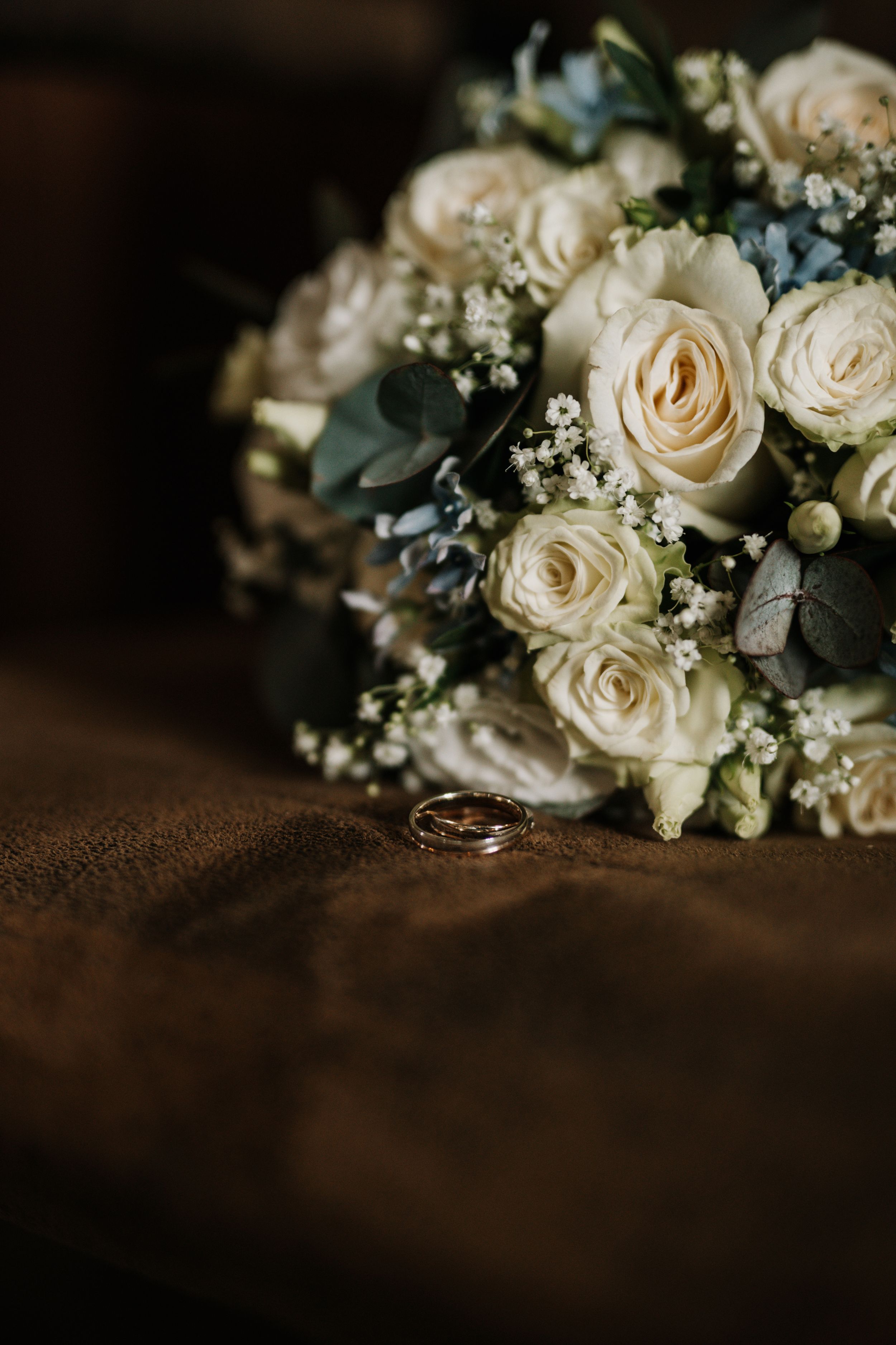 A delicate white rose wedding bouquet with baby's breath and greenery beside wedding rings.