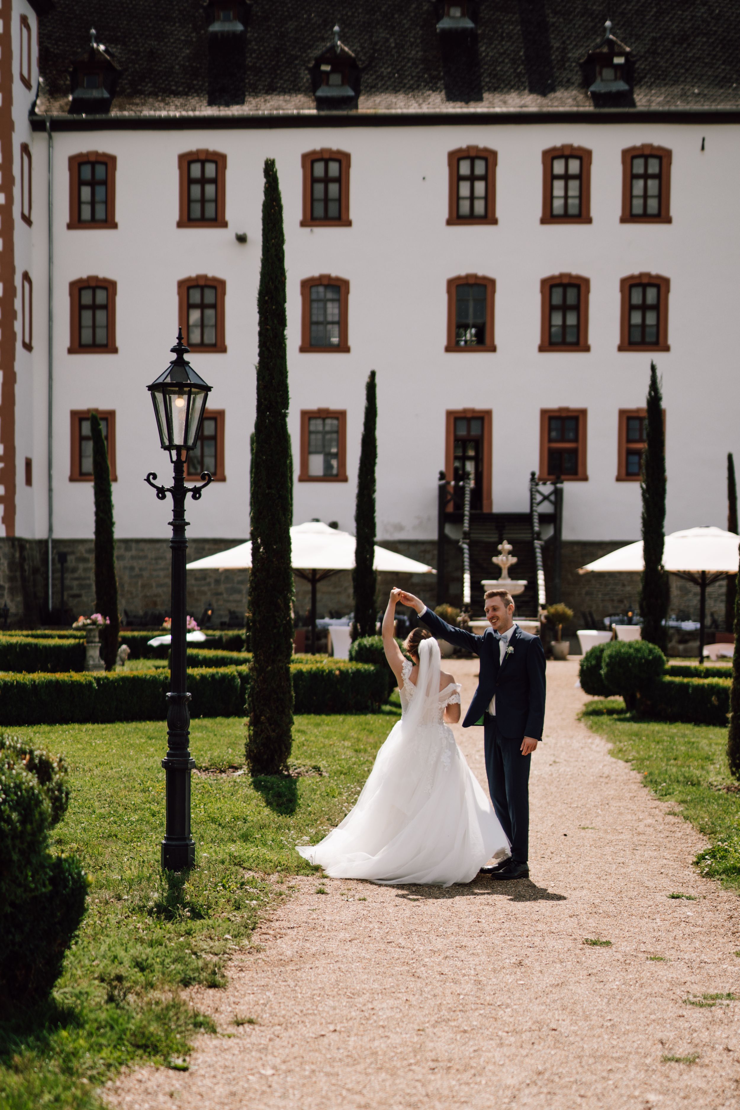 A wedding couple walks along a gravel path beside a grand white building with cypress trees.