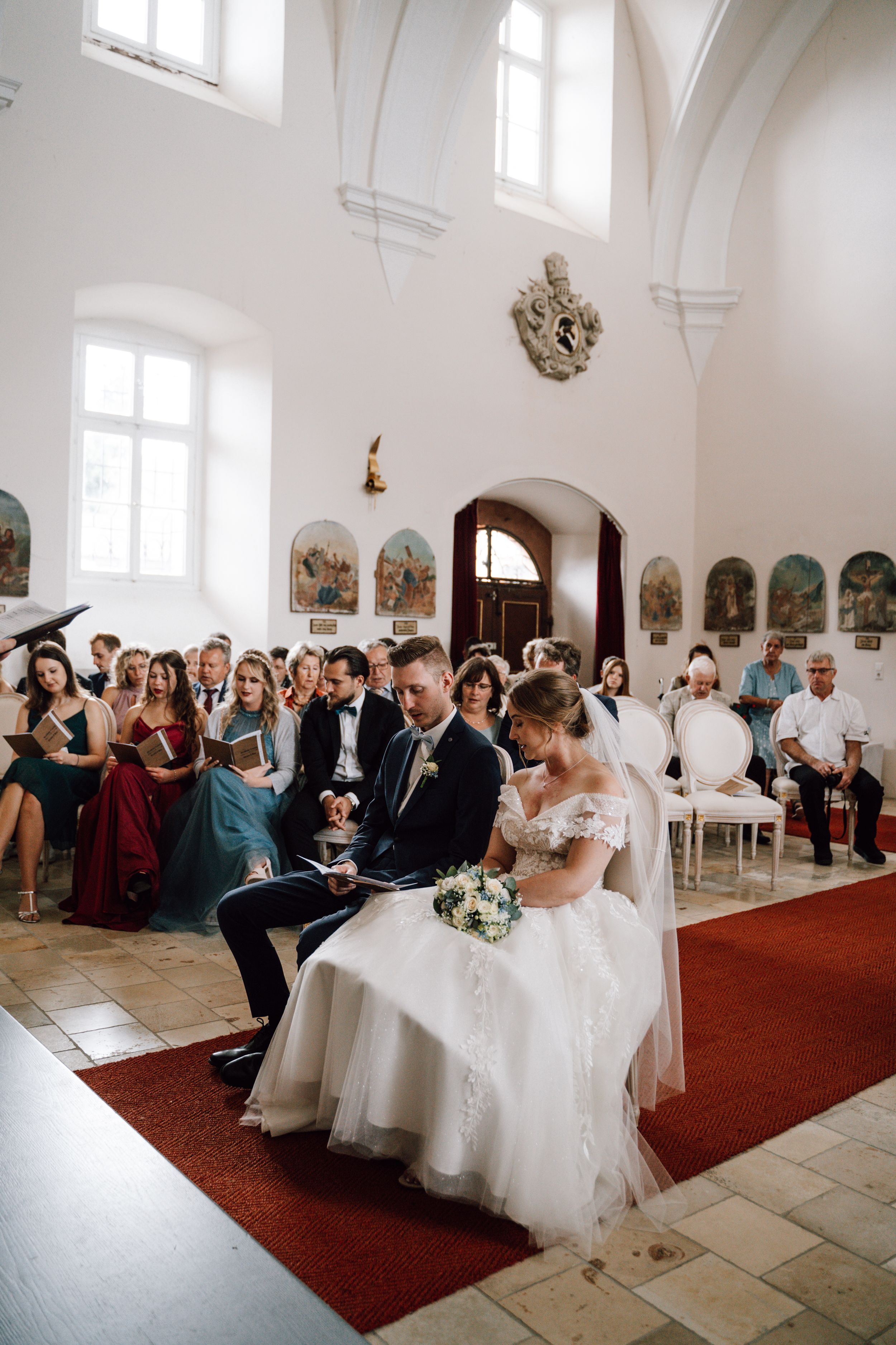 A wedding ceremony in a bright white church with guests seated on wooden chairs.