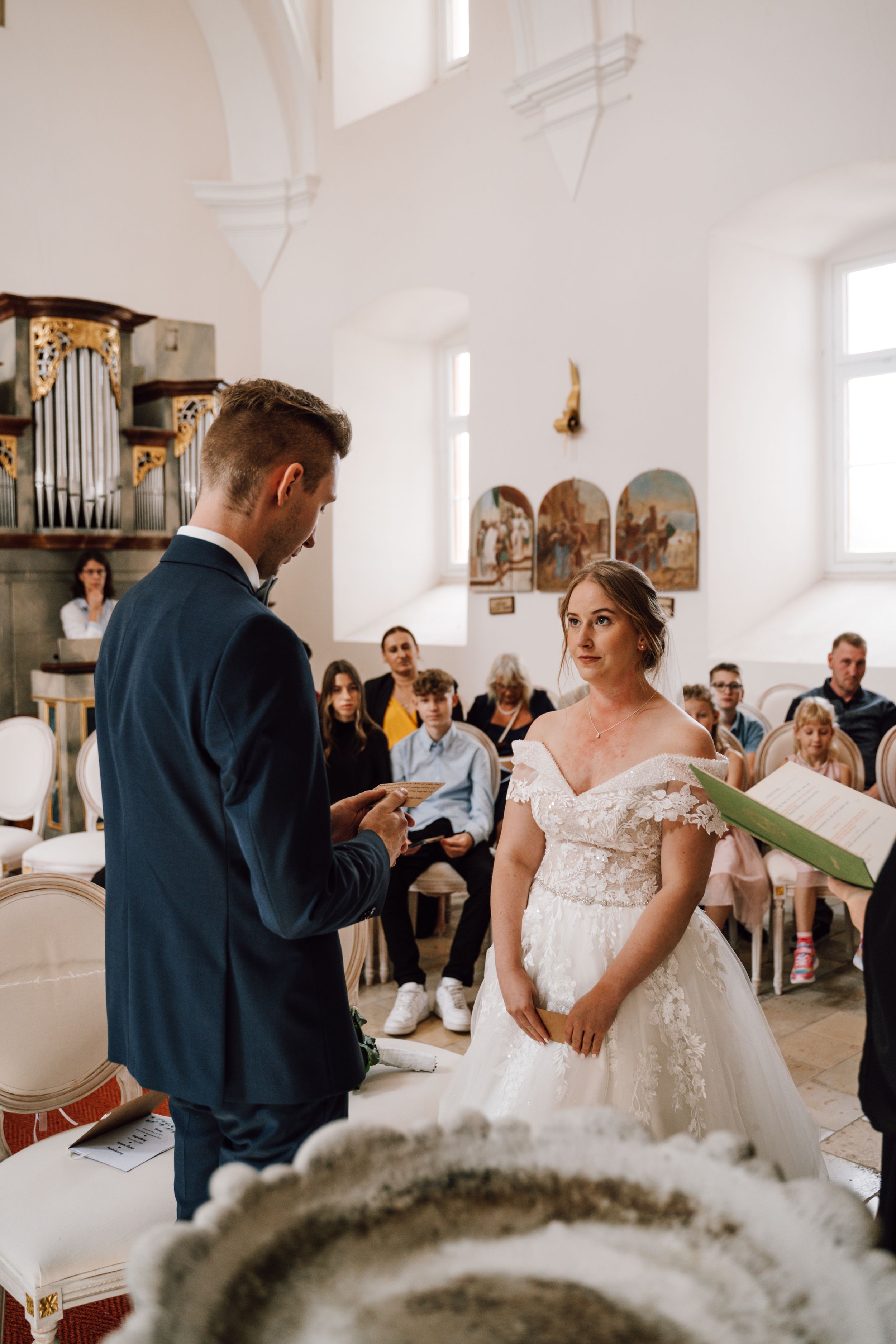 A wedding ceremony taking place in a beautiful white church with high ceilings and religious artwork.