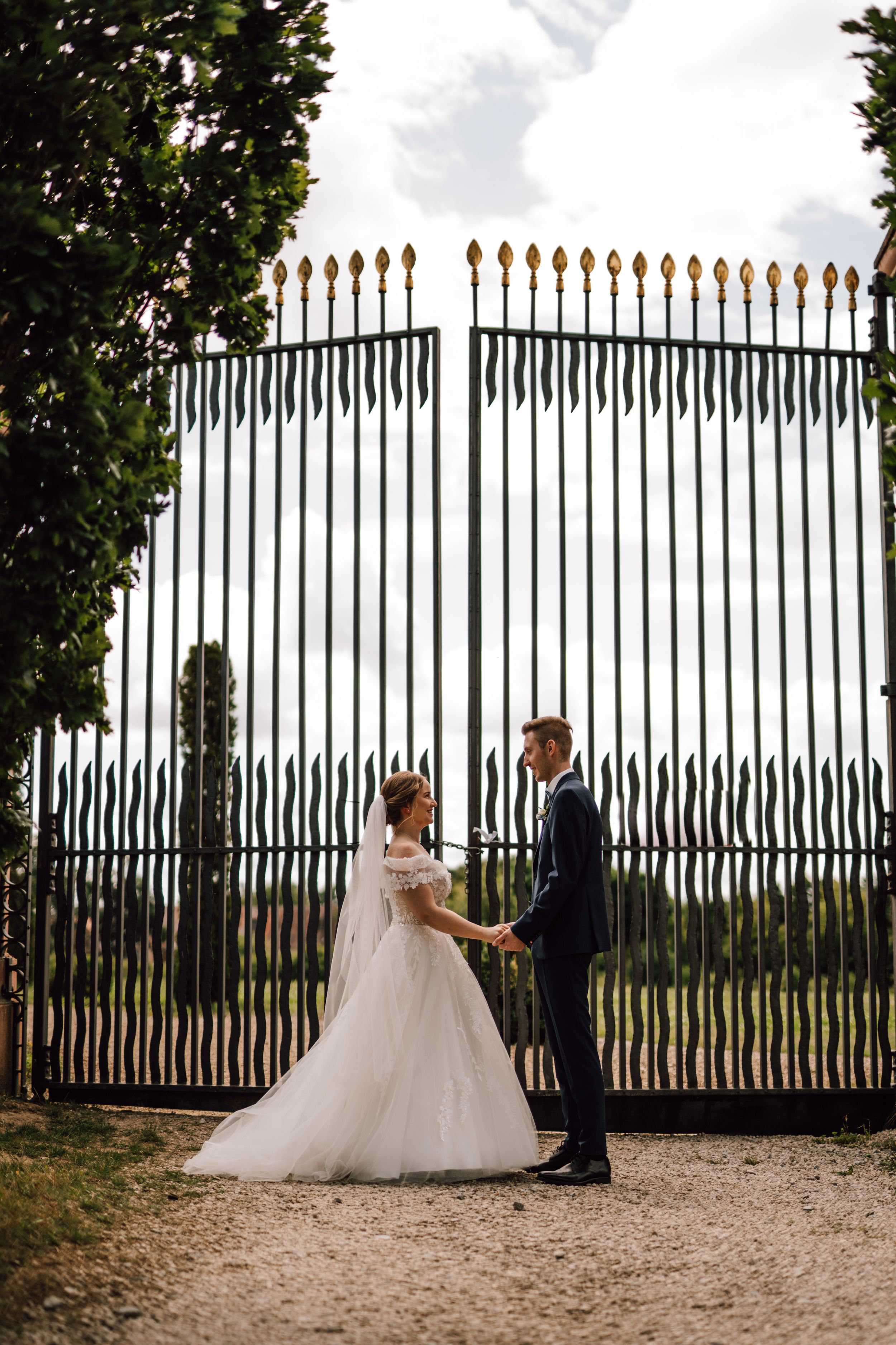 A wedding couple poses by an ornate iron fence with golden spikes against green foliage.