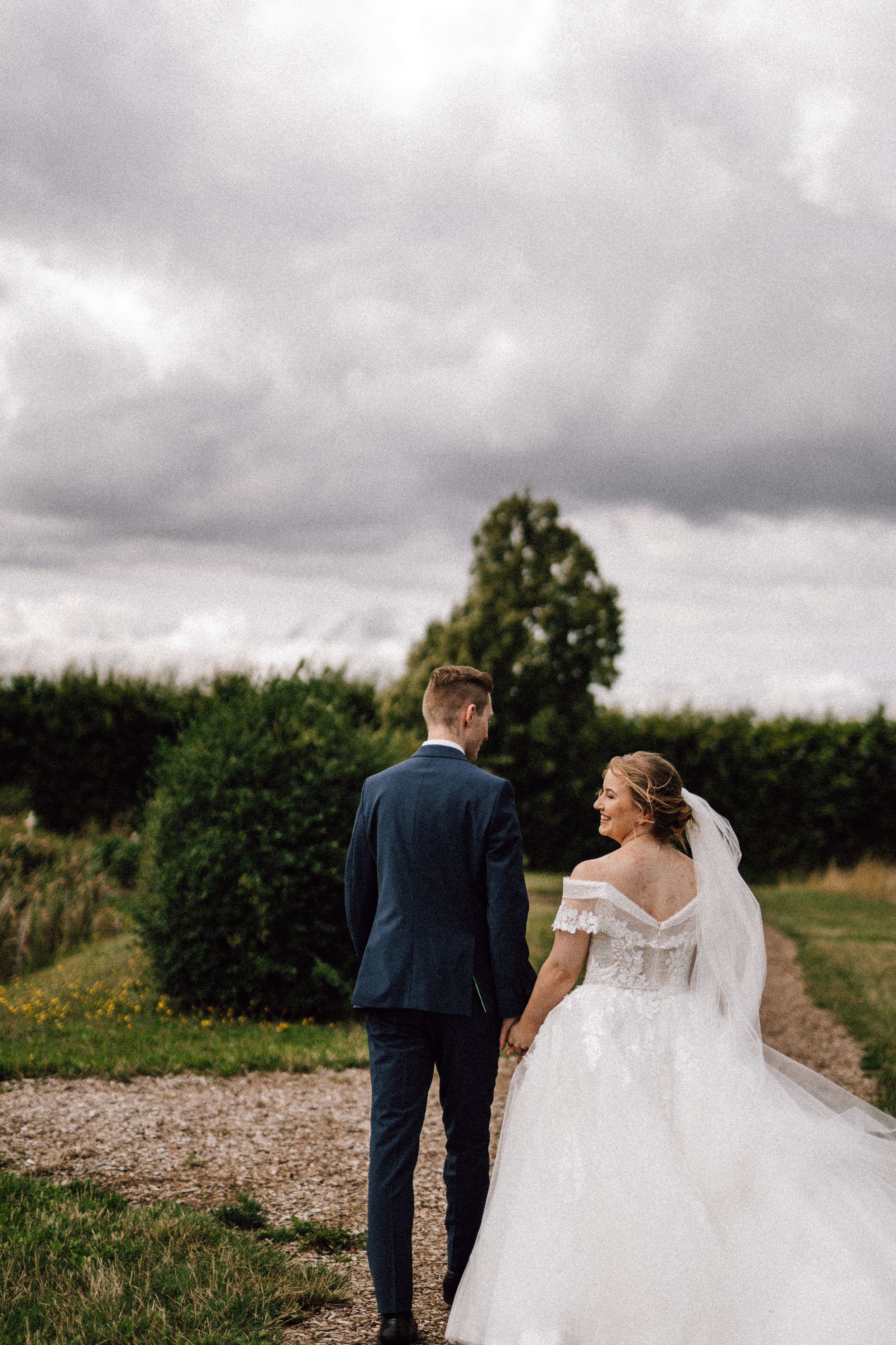 A bride and groom walk hand in hand on a path under dramatic cloudy skies surrounded by greenery.