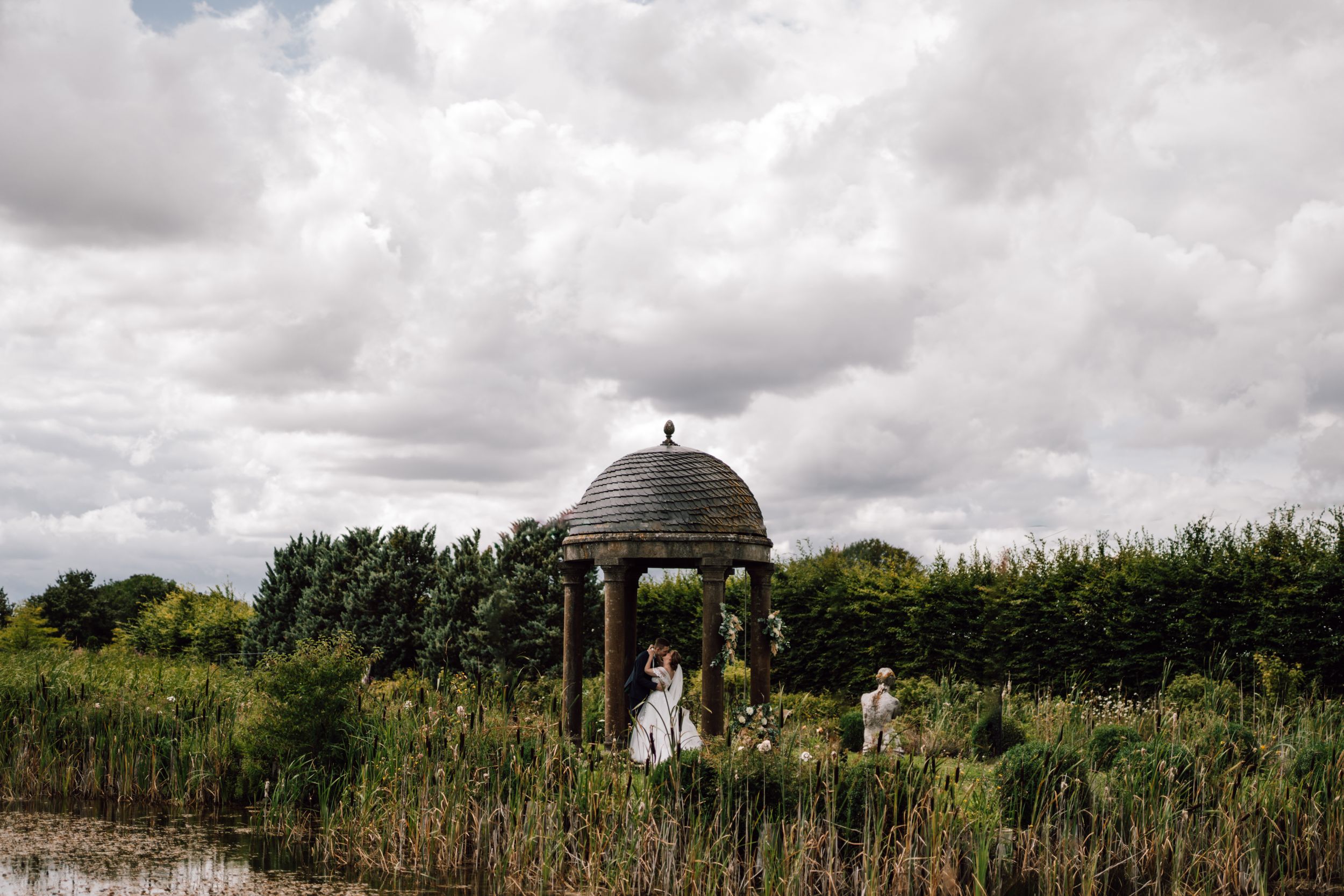 A wedding ceremony takes place under a domed gazebo surrounded by gardens and hedges.