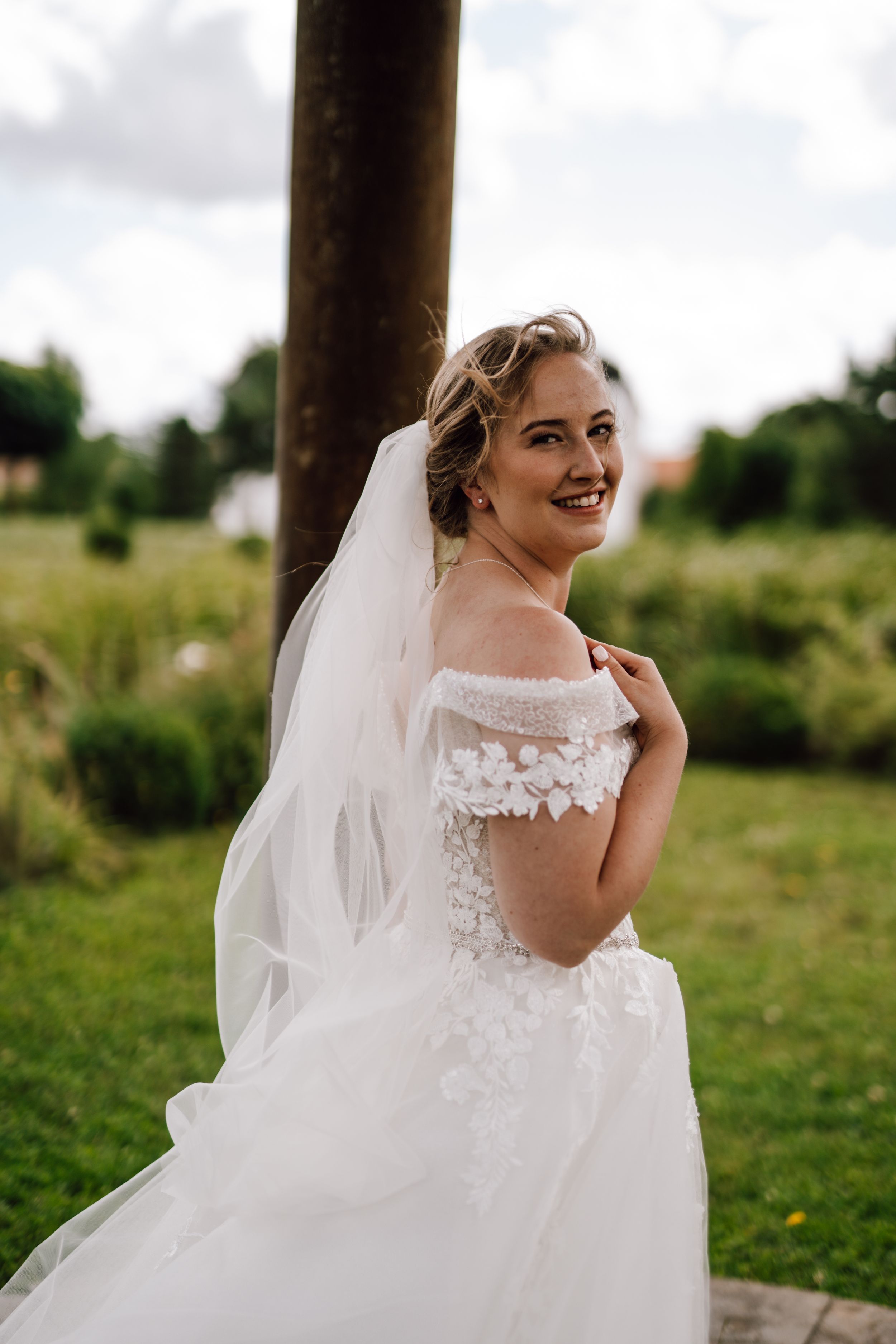 A bride in an off-shoulder lace wedding dress poses outdoors with a flowing veil.