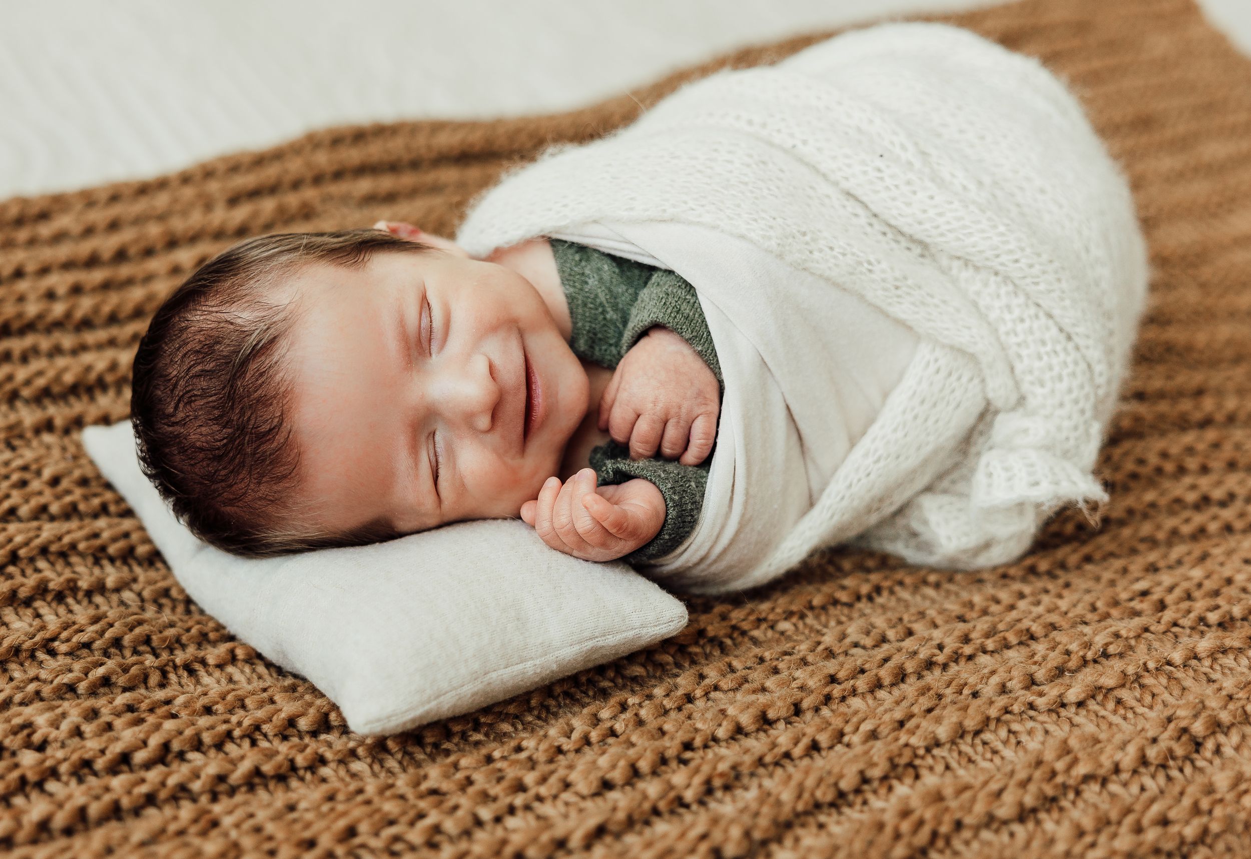 A sleeping infant swaddled in a white blanket lies peacefully on a brown knit blanket.