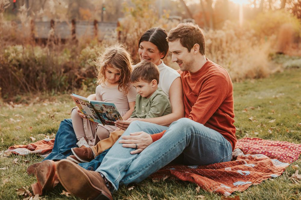 Family enjoying reading together on a red blanket in a park during golden hour.