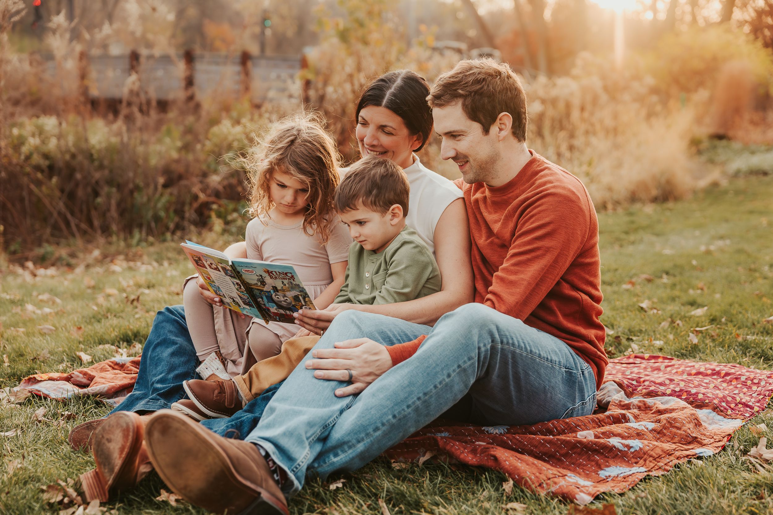 Family enjoying reading together on a red blanket in a park during golden hour.