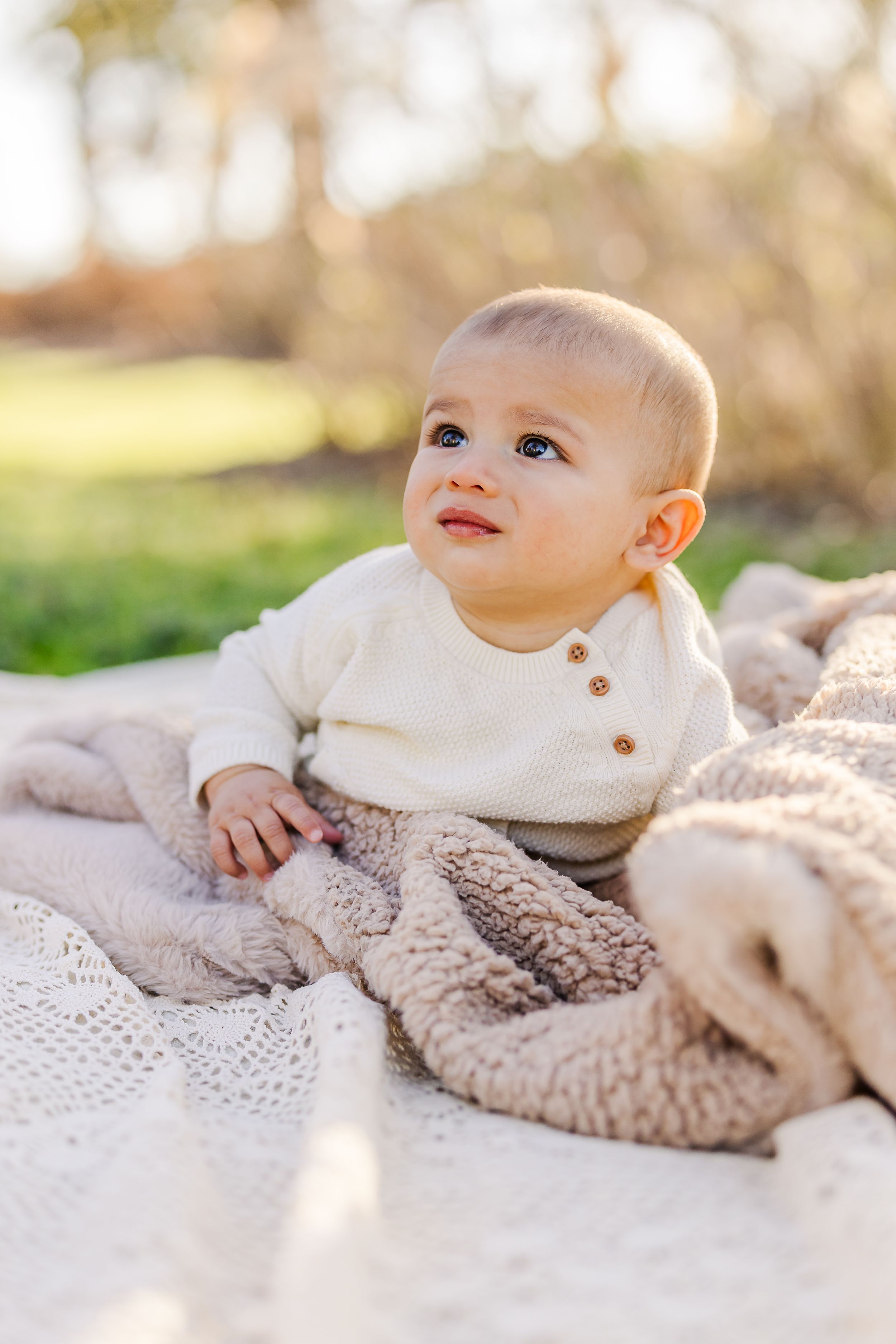 Baby in cream sweater lies on cozy blanket outdoors during golden hour photo session.