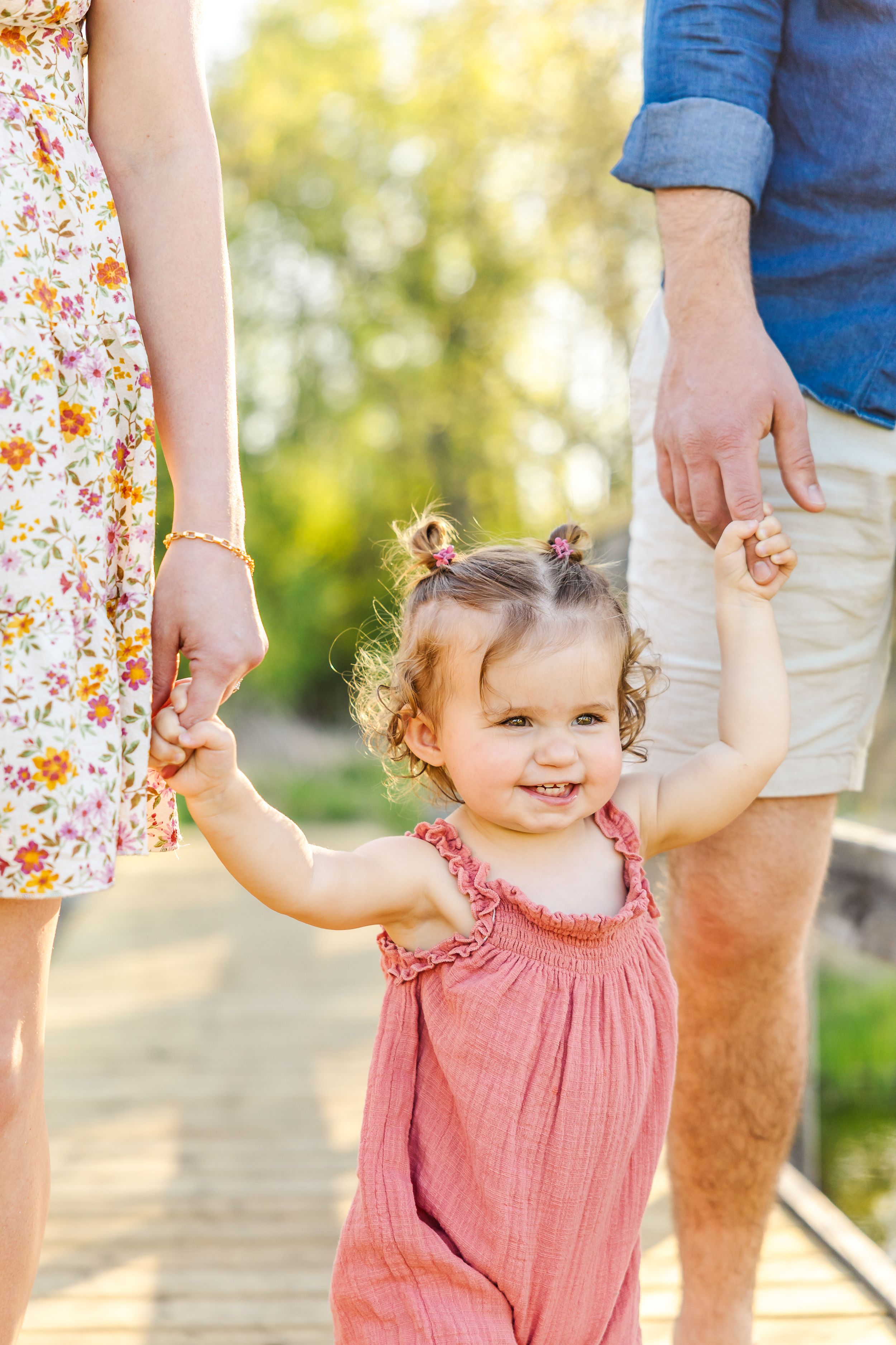 A small child in a pink dress holds hands with parents while walking outdoors on a sunny day with a blurred background.