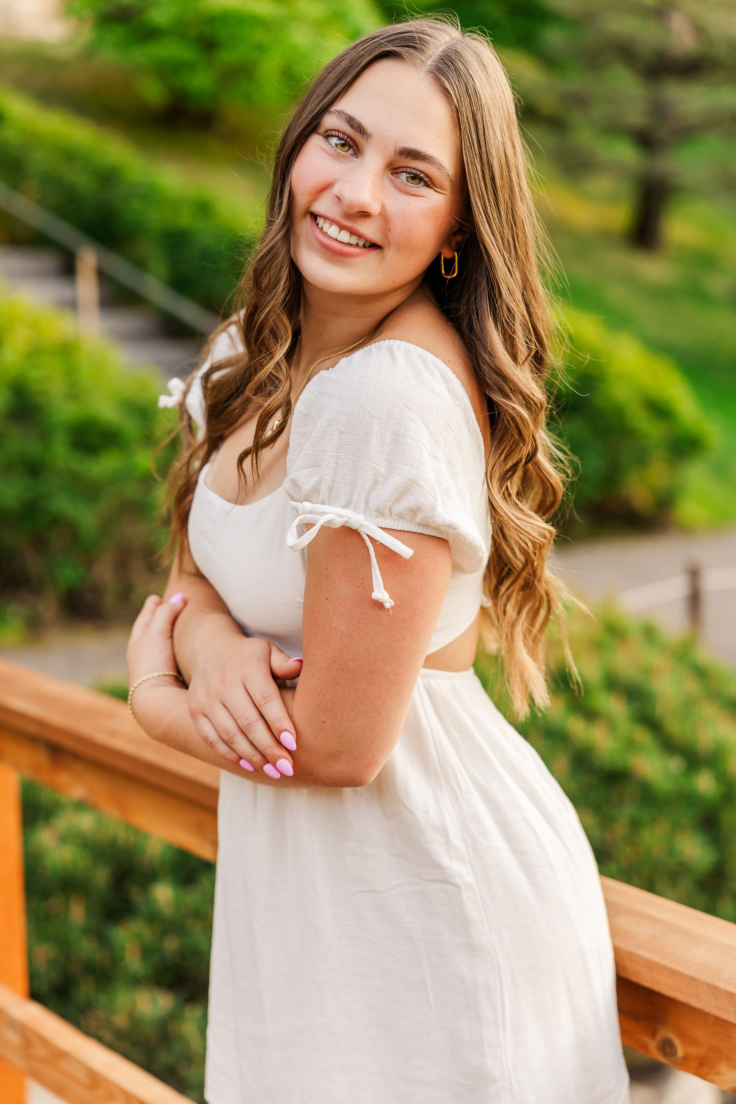 Person wearing a white summer dress with puff sleeves poses on a wooden deck railing with green foliage in background.