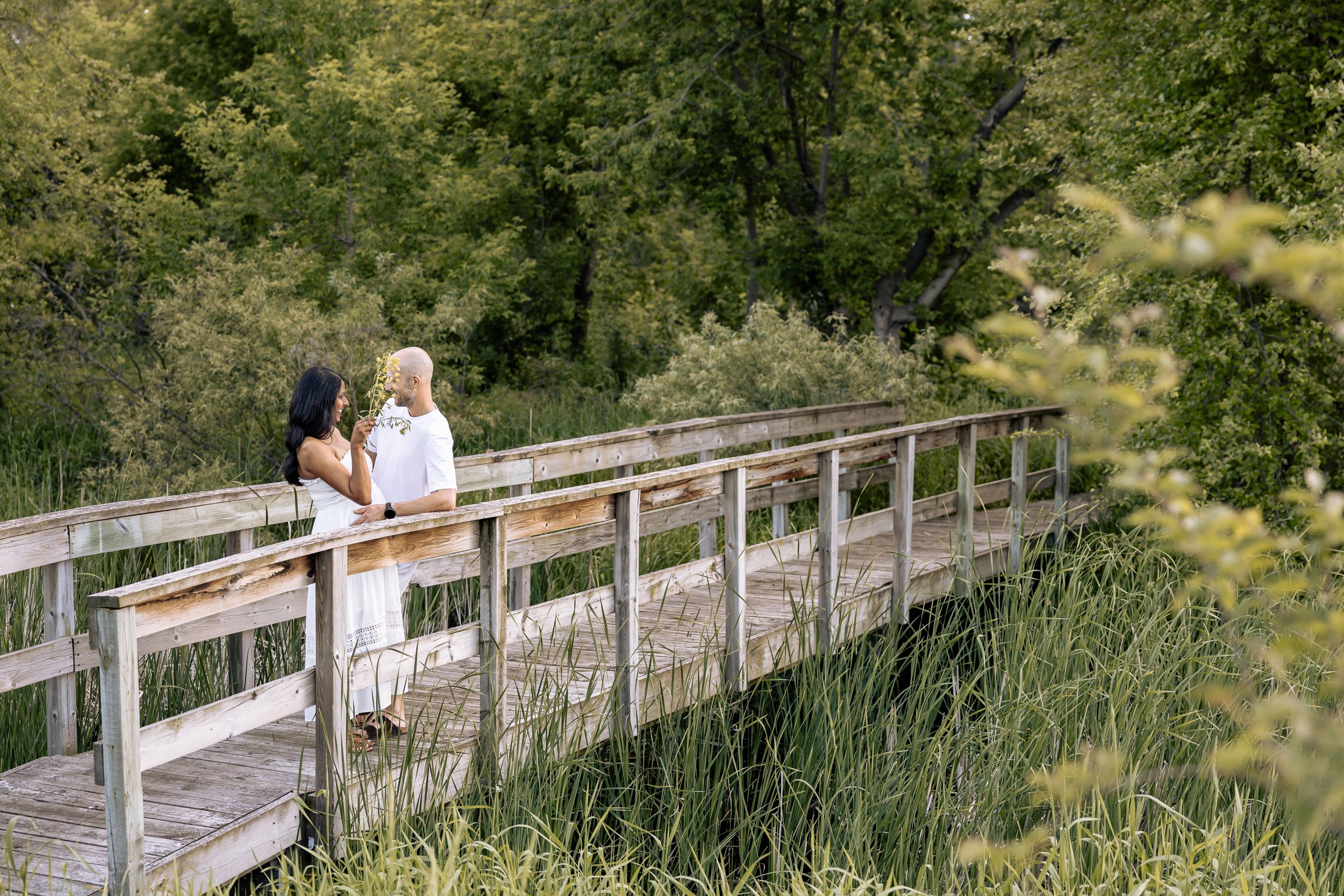 Two people standing together on a wooden boardwalk surrounded by lush greenery.