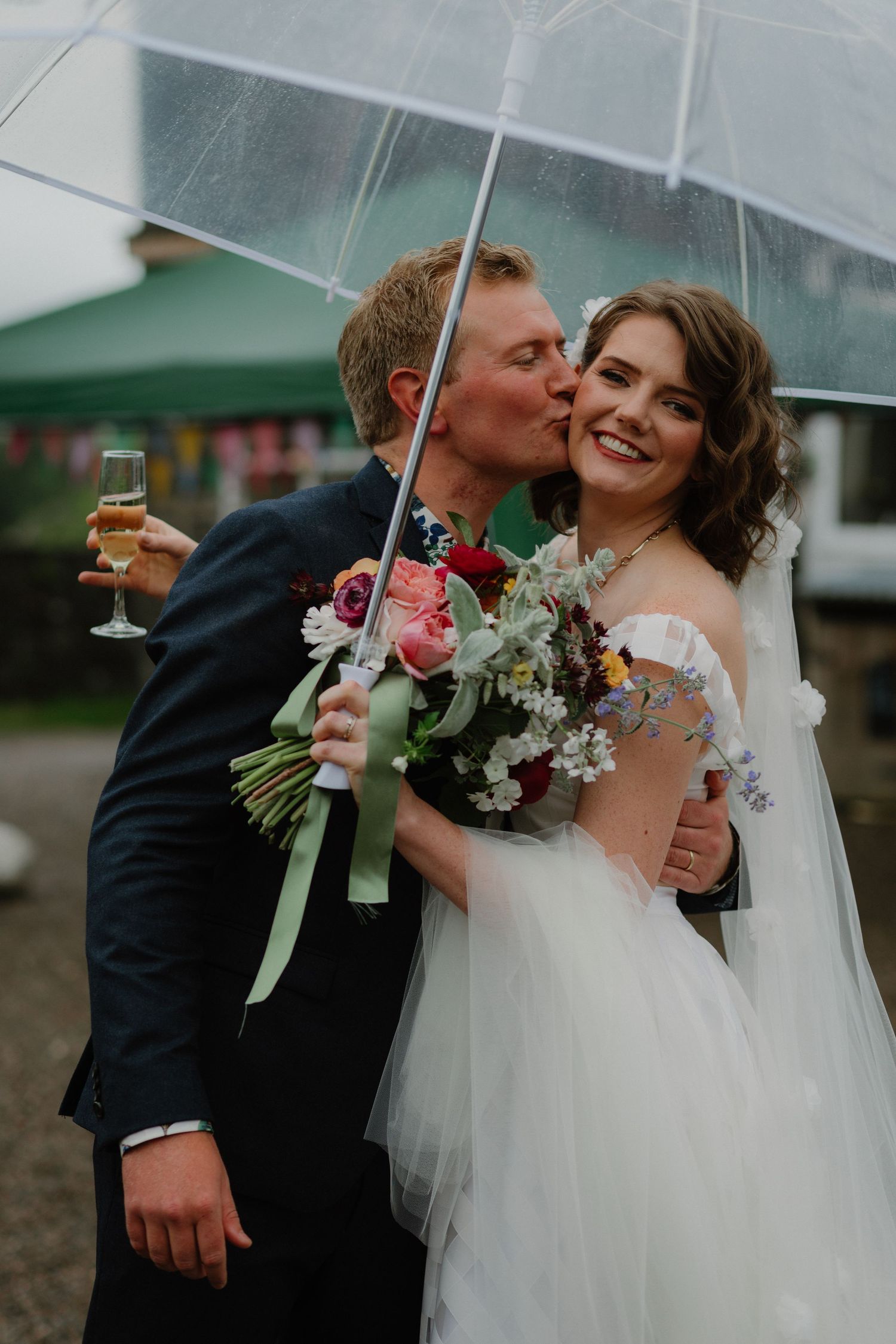 A couple shares a joyful moment under a clear umbrella while holding champagne and a bouquet.