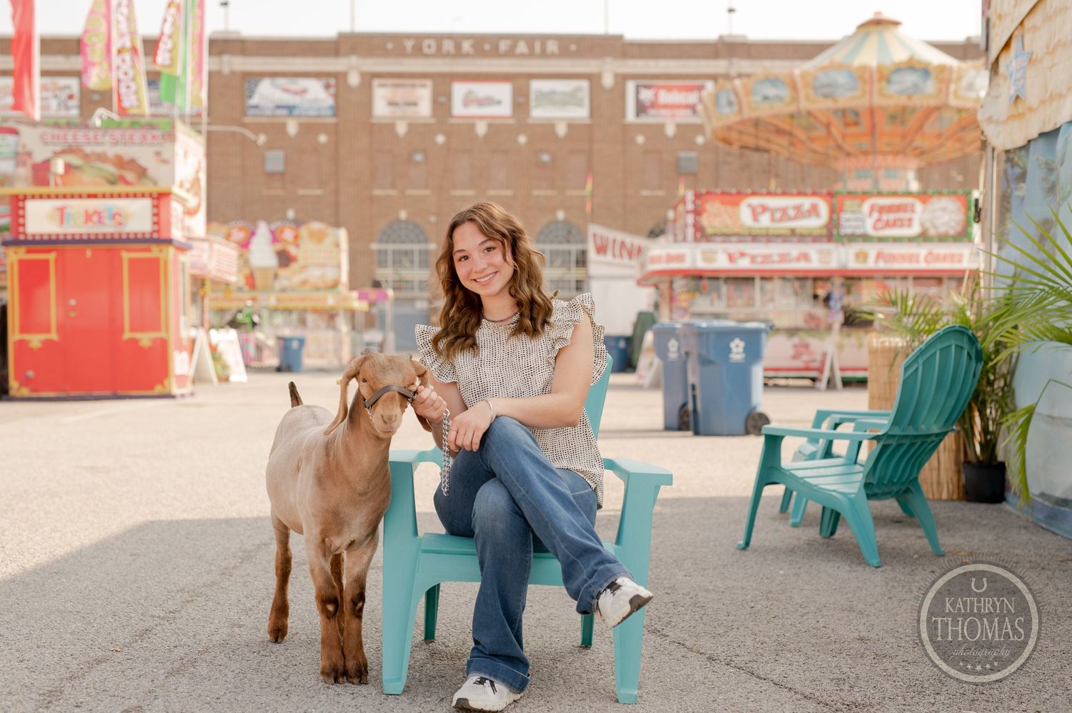 Brown goat stands with owner at fairgrounds near red food stands and carnival attractions.