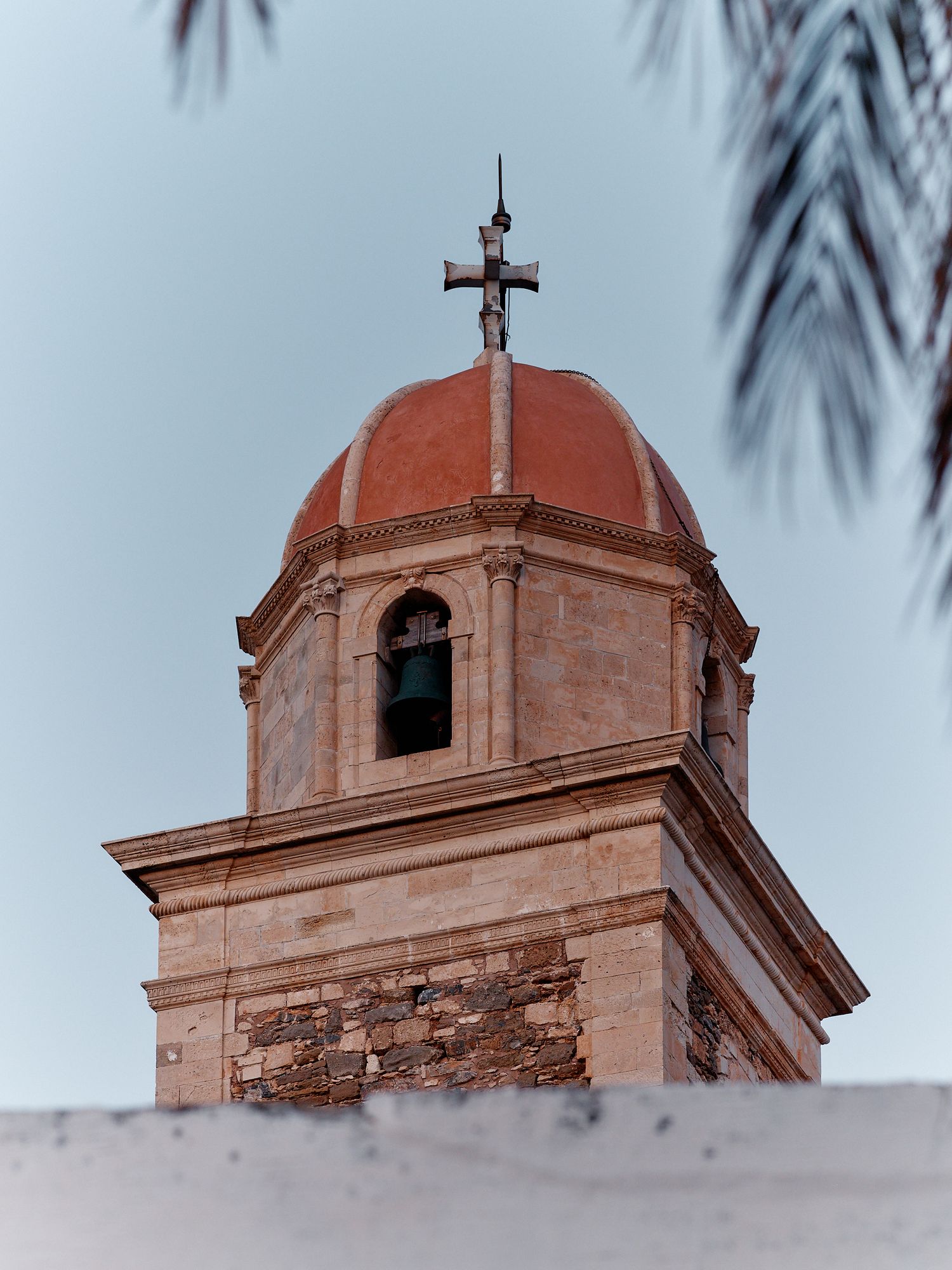 Pink-hued bell tower with cross sits atop a historic church against palm fronds at sunset.